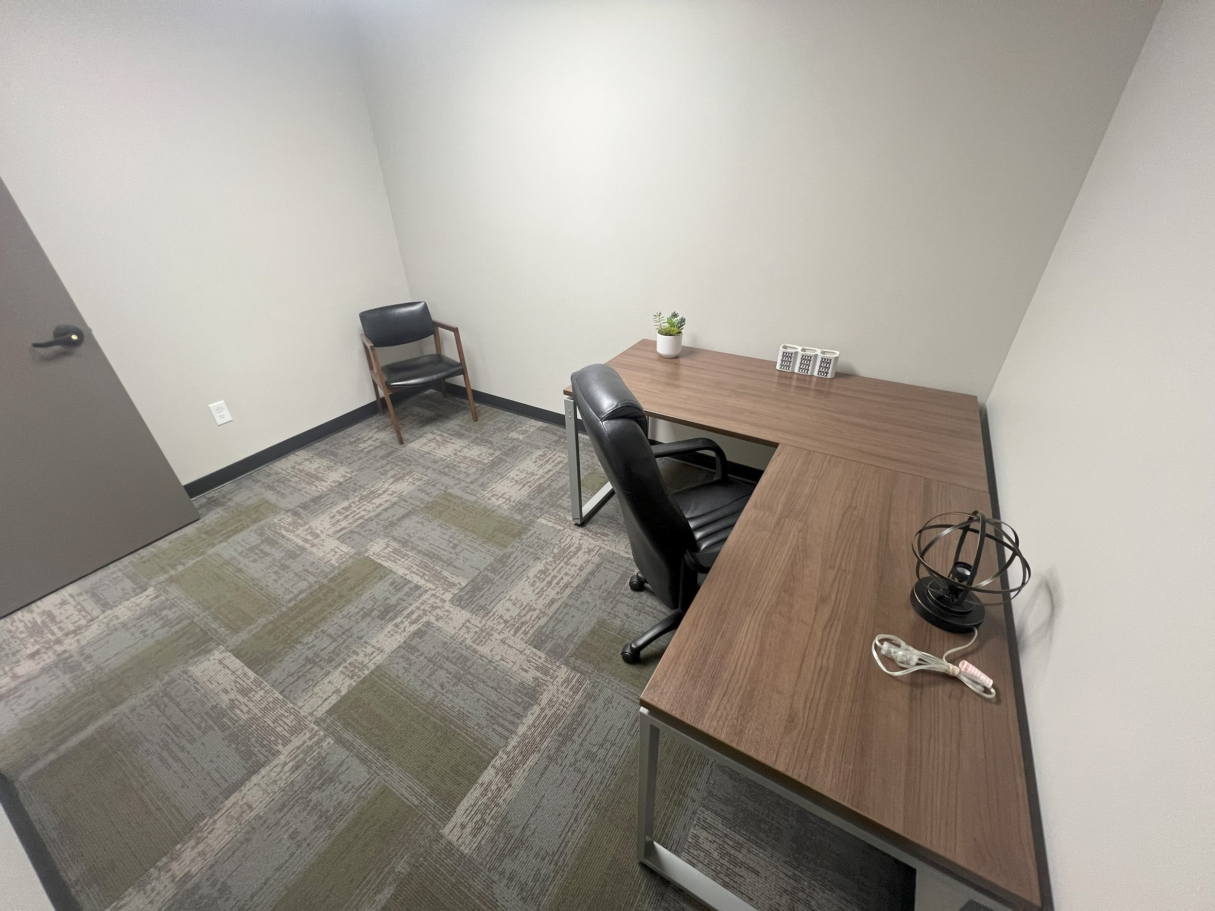 Small office room with a brown L-shaped desk, a black office chair, a small black chair, a potted plant, a white power strip, a black wireframe lamp, and a white Ethernet cable.