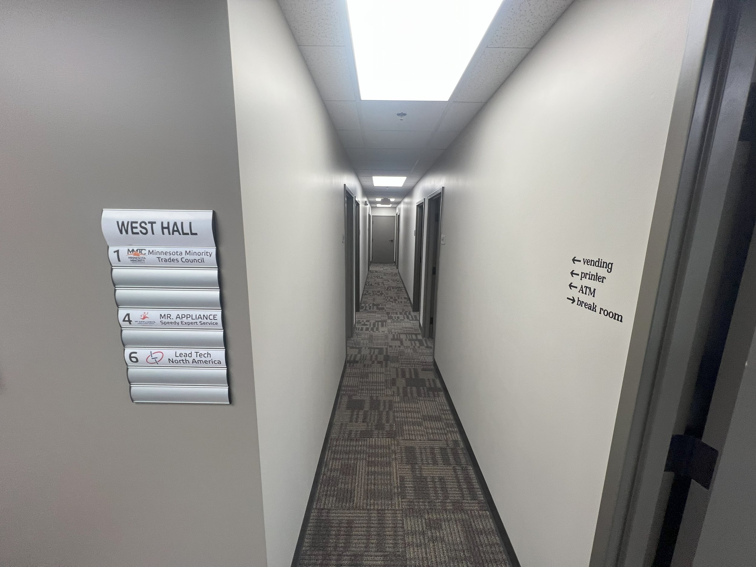 A hallway with doorways on both sides, a ceiling with fluorescent lighting, and signs indicating offices and facilities at the end of the hallway.
