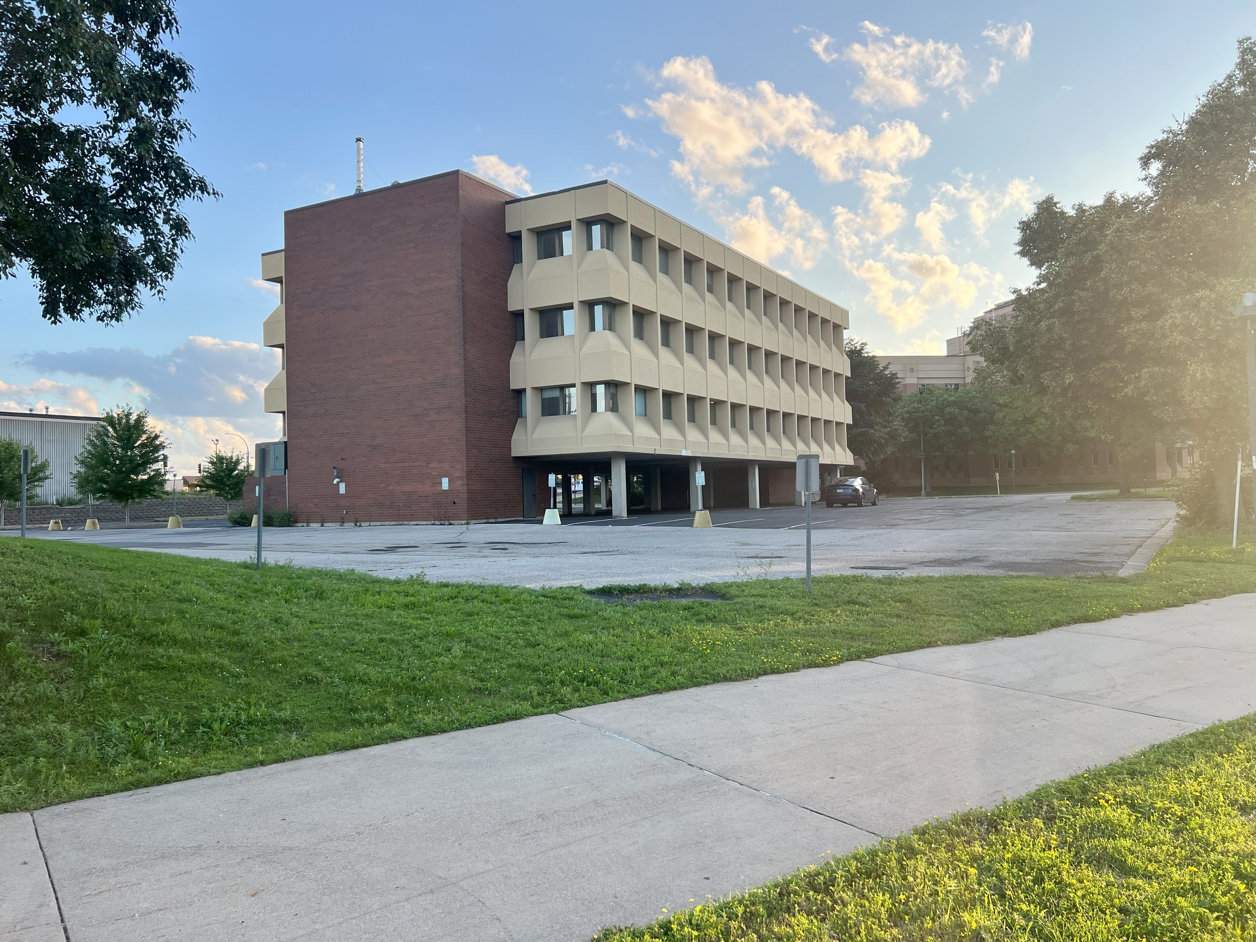 A multi-story parking garage supported by concrete pillars with a small entrance on the ground level, located on a paved lot with a single car parked on the far side. The building has a distinctive design with curved balcony-like structures and a bri