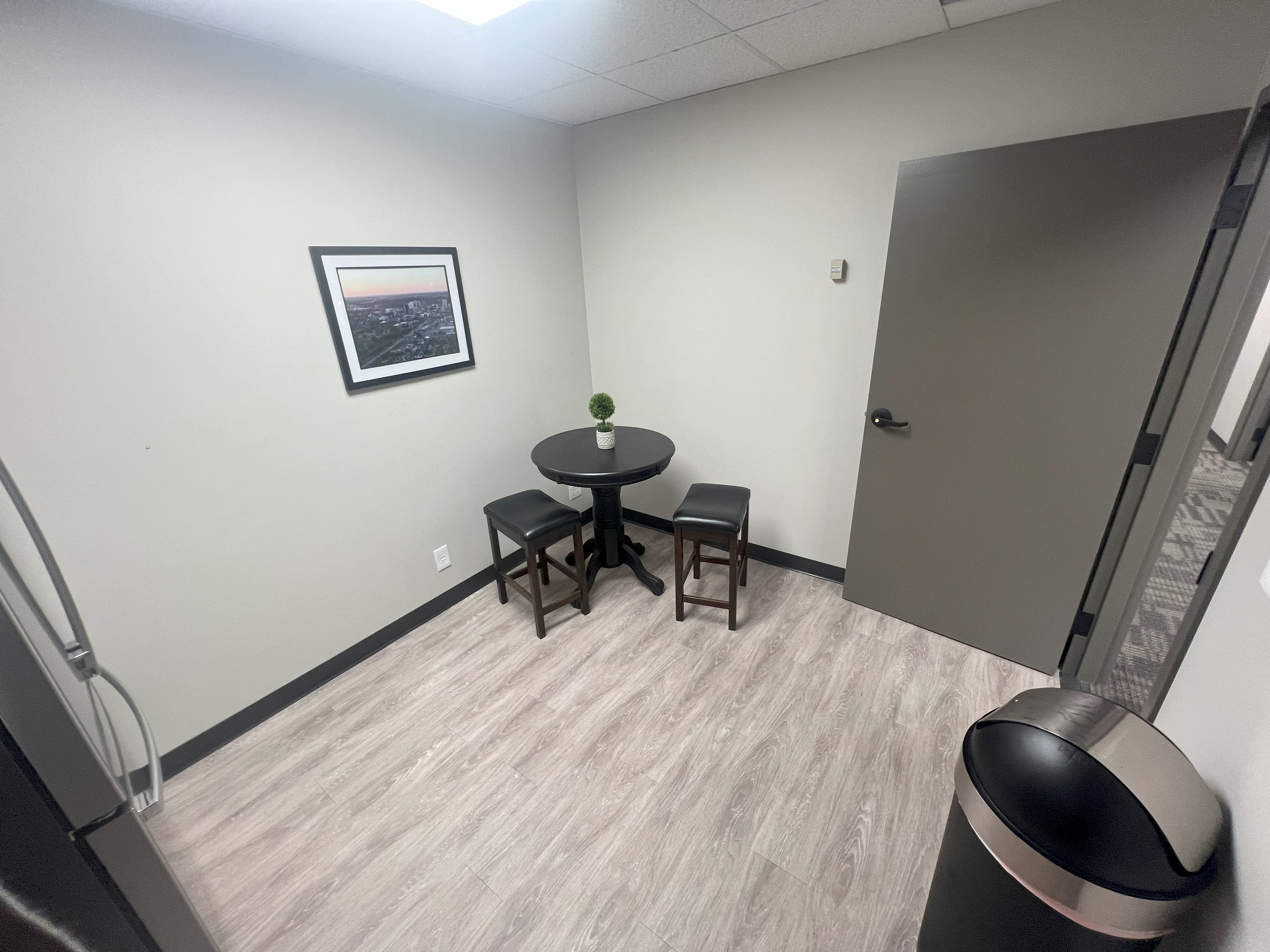 Small office corner with a black round table, two black stools, a potted plant, wall art, gray door, and a trash can.