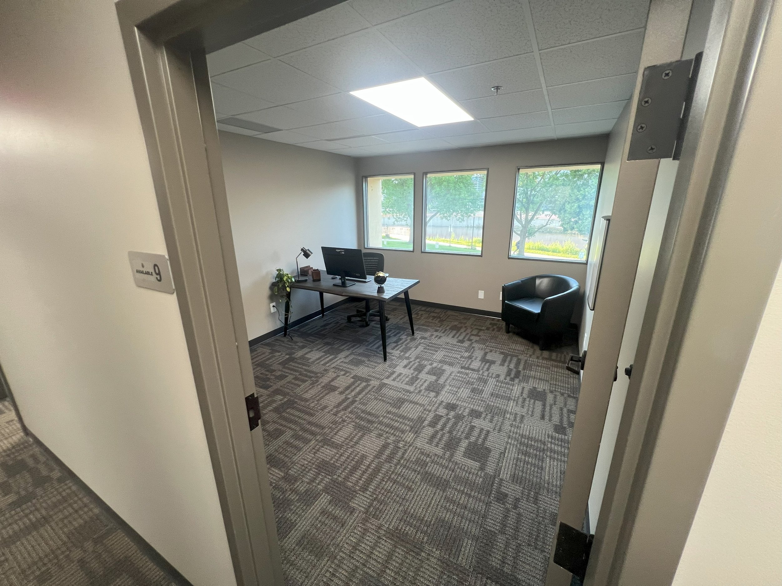 Empty office room with a desk, computer, and a black armchair near windows with trees outside.