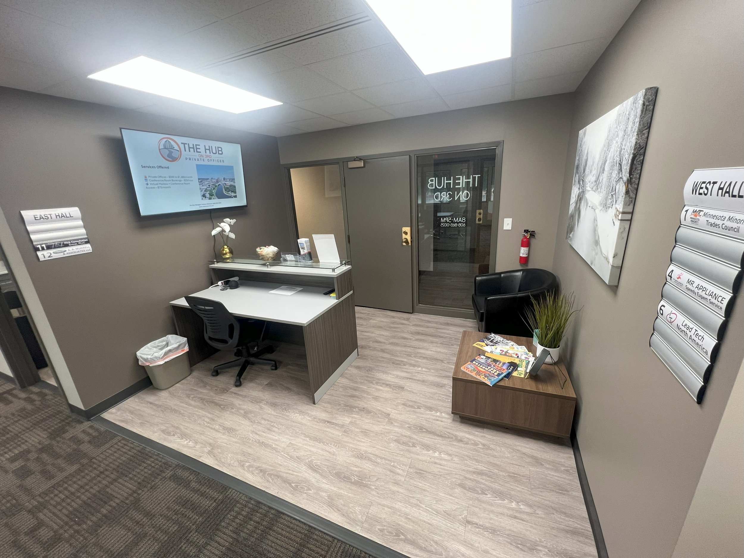 Office reception area with a desk, black chair, side table with magazines and plant, wall-mounted screen displaying information, and various signs on the walls.