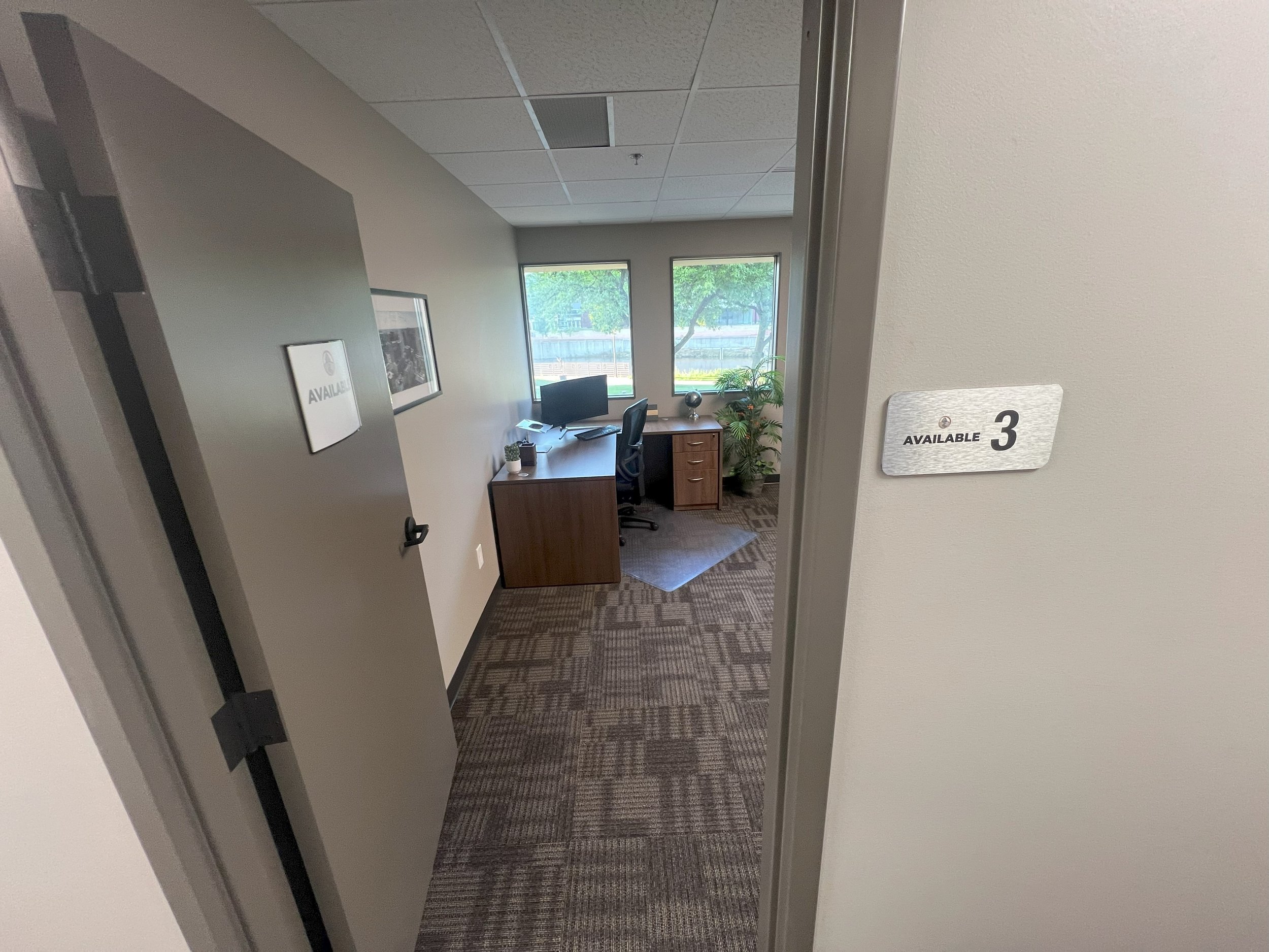 Office room with a desk, computer, chair, potted plant, and windows showing trees outside. Sign on wall reads 'AVAILABLE 3'.