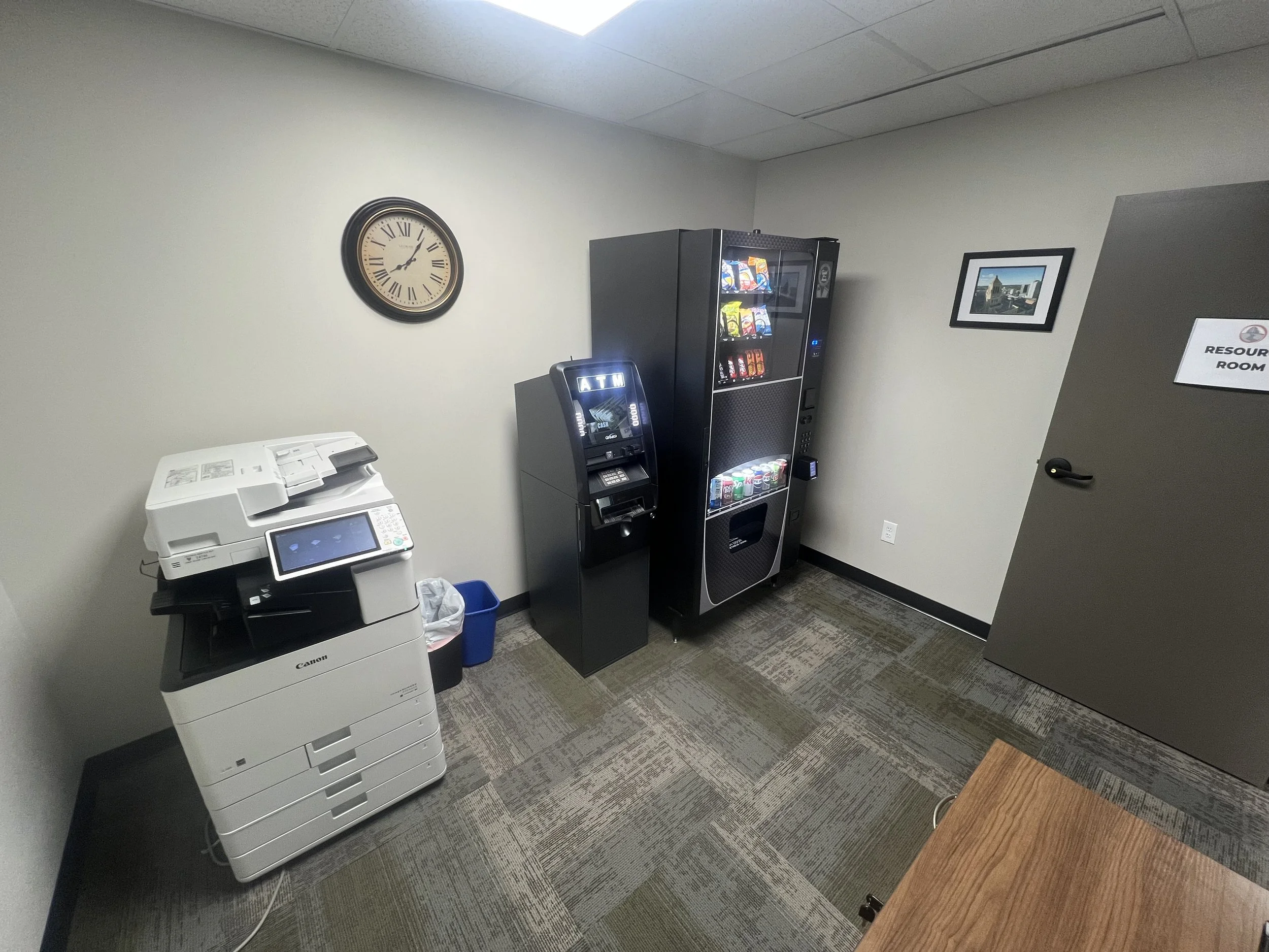 Office break room with a wall clock, a Xerox multifunction printer, a vending machine, a cashless ATM, and a resource room door.