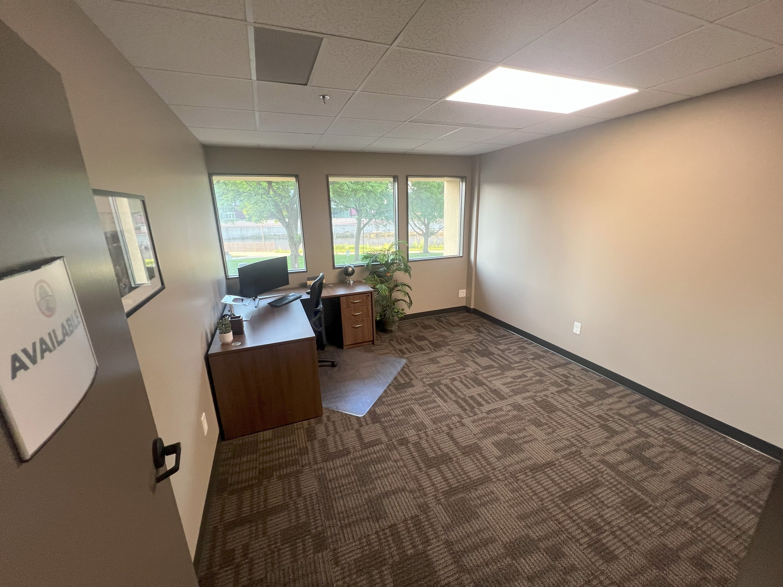 Empty office room with three windows, a desk with a computer, a chair, a small filing cabinet, a potted plant, and a framed sign on the wall.