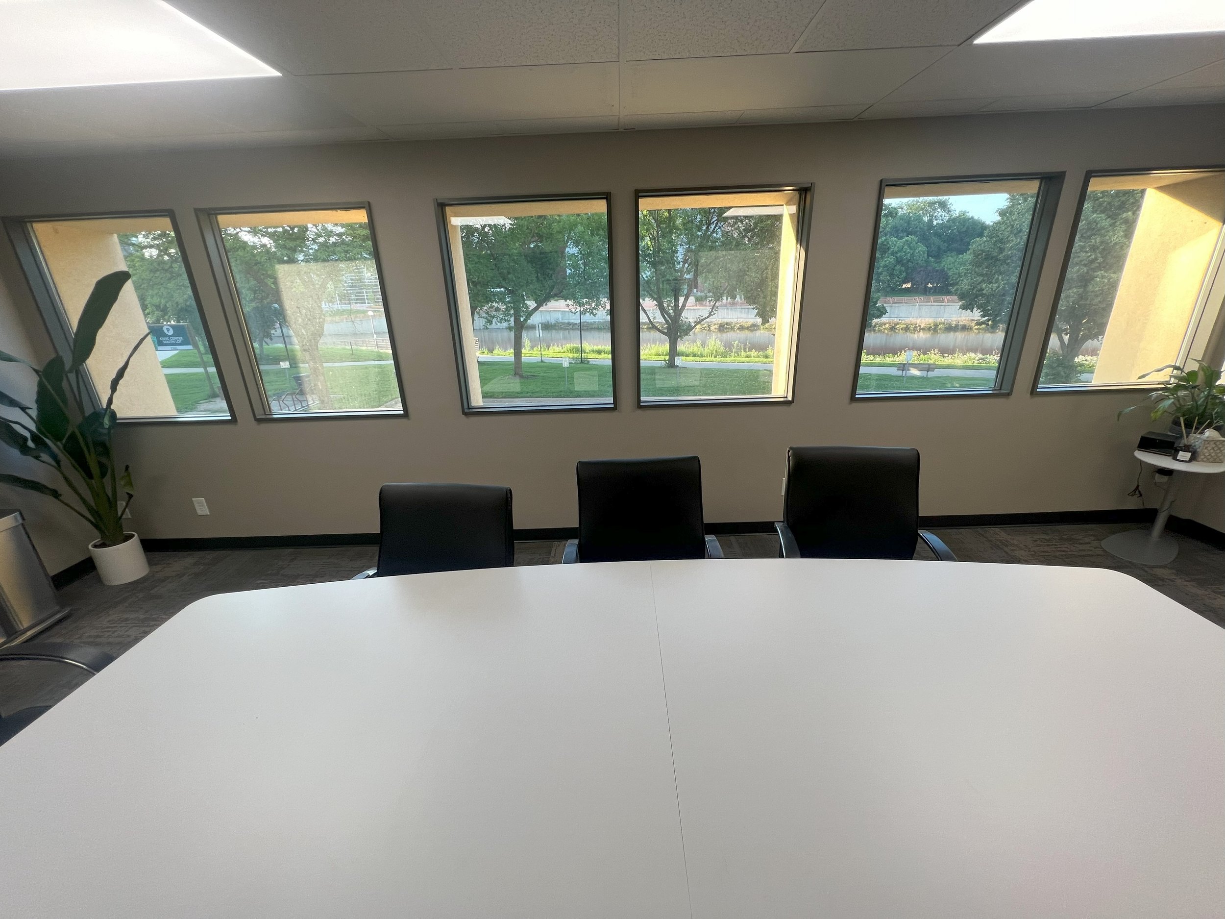 Empty conference room with a large white table, three black chairs, a potted plant, and four windows showing a view of trees and a lake outside.