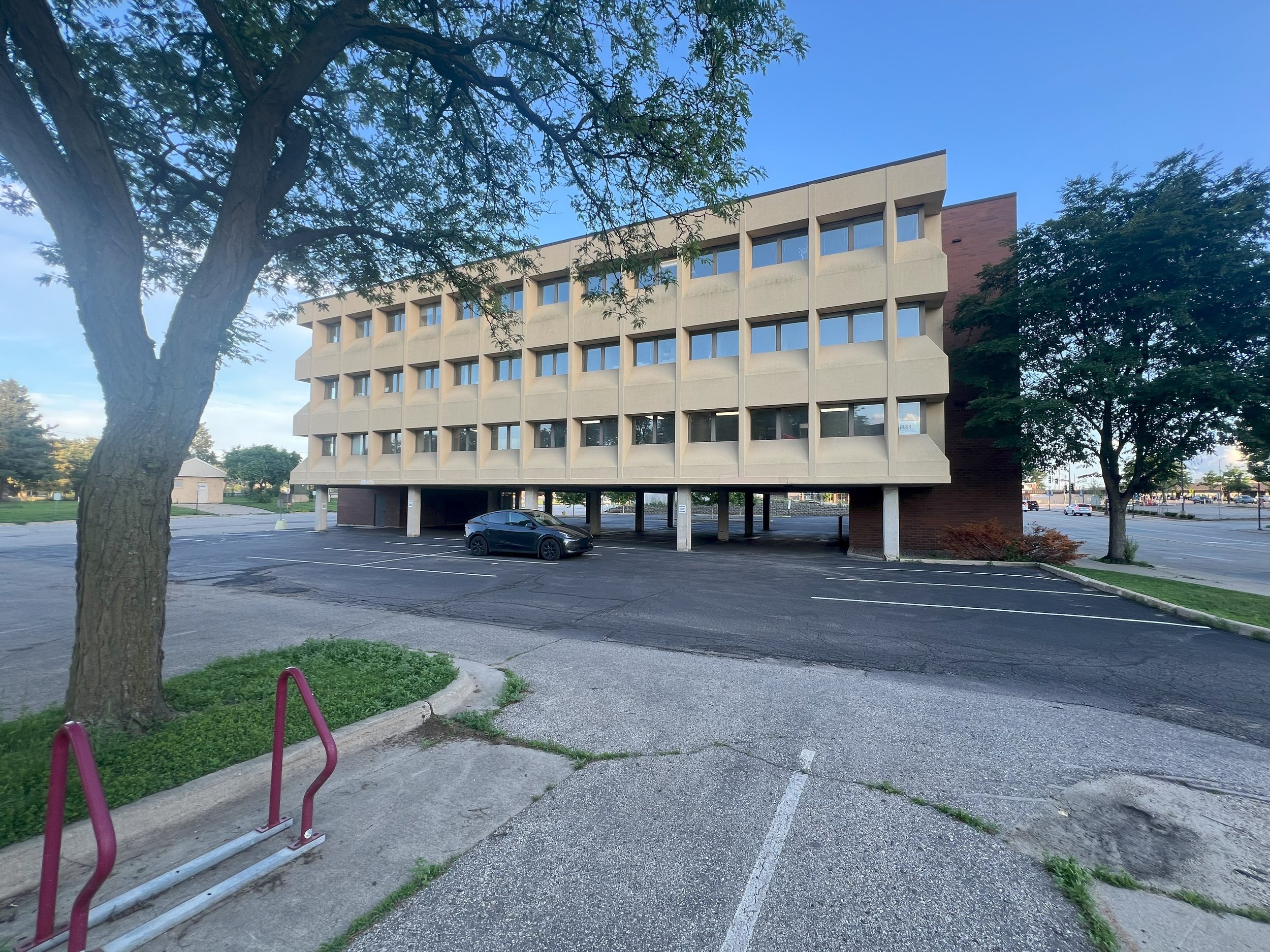 A multi-story office building with a parking area underneath, surrounded by trees, blue sky, and an empty parking lot in front.