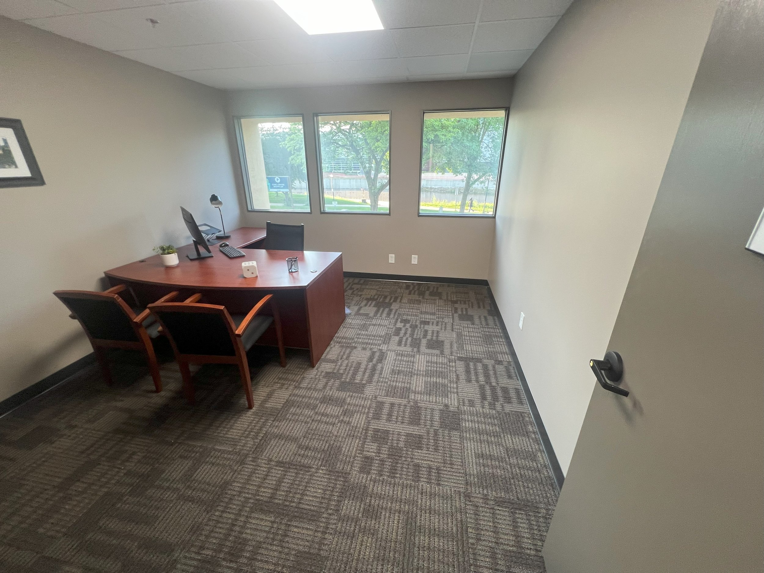 An empty office with a wooden desk, two black chairs in front of the desk, a black office chair behind it, a computer monitor, a desk lamp, and a small potted plant. There are three large windows showing trees outside.