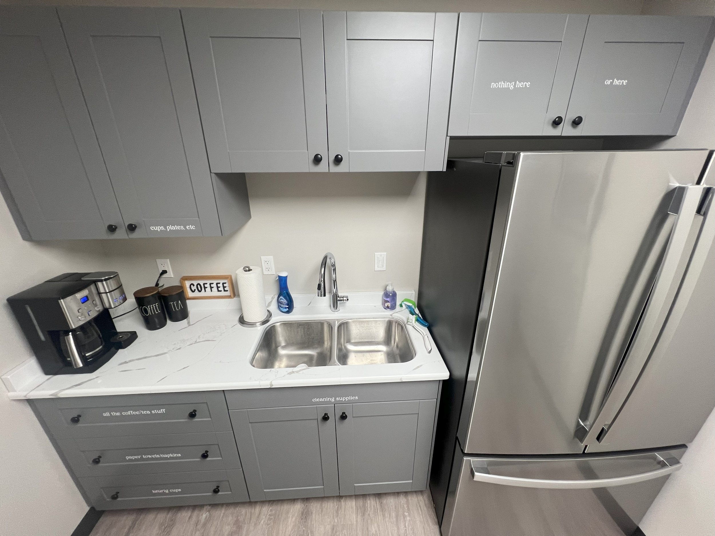 Small kitchen area with gray cabinets, stainless steel refrigerator, white marble countertop, and various kitchen items including coffee maker, mugs, soap dispenser, and dish soap.