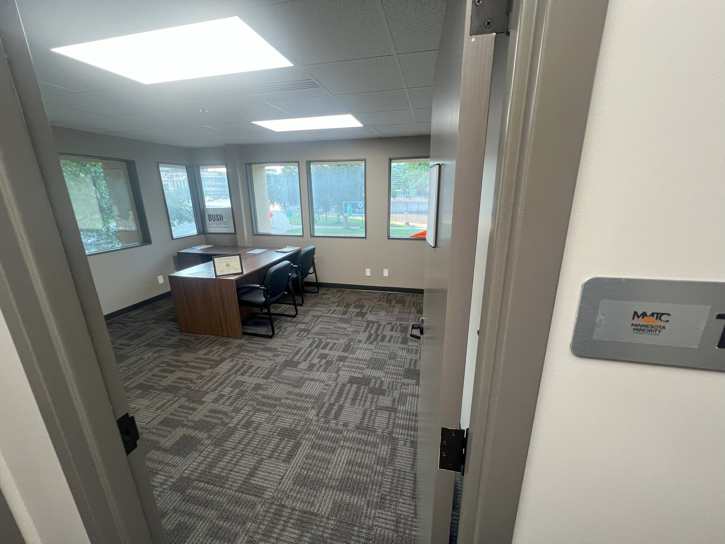 Empty office with a desk and three chairs, windows with a view outside, and a sign that reads "BUSH" visible through the windows.