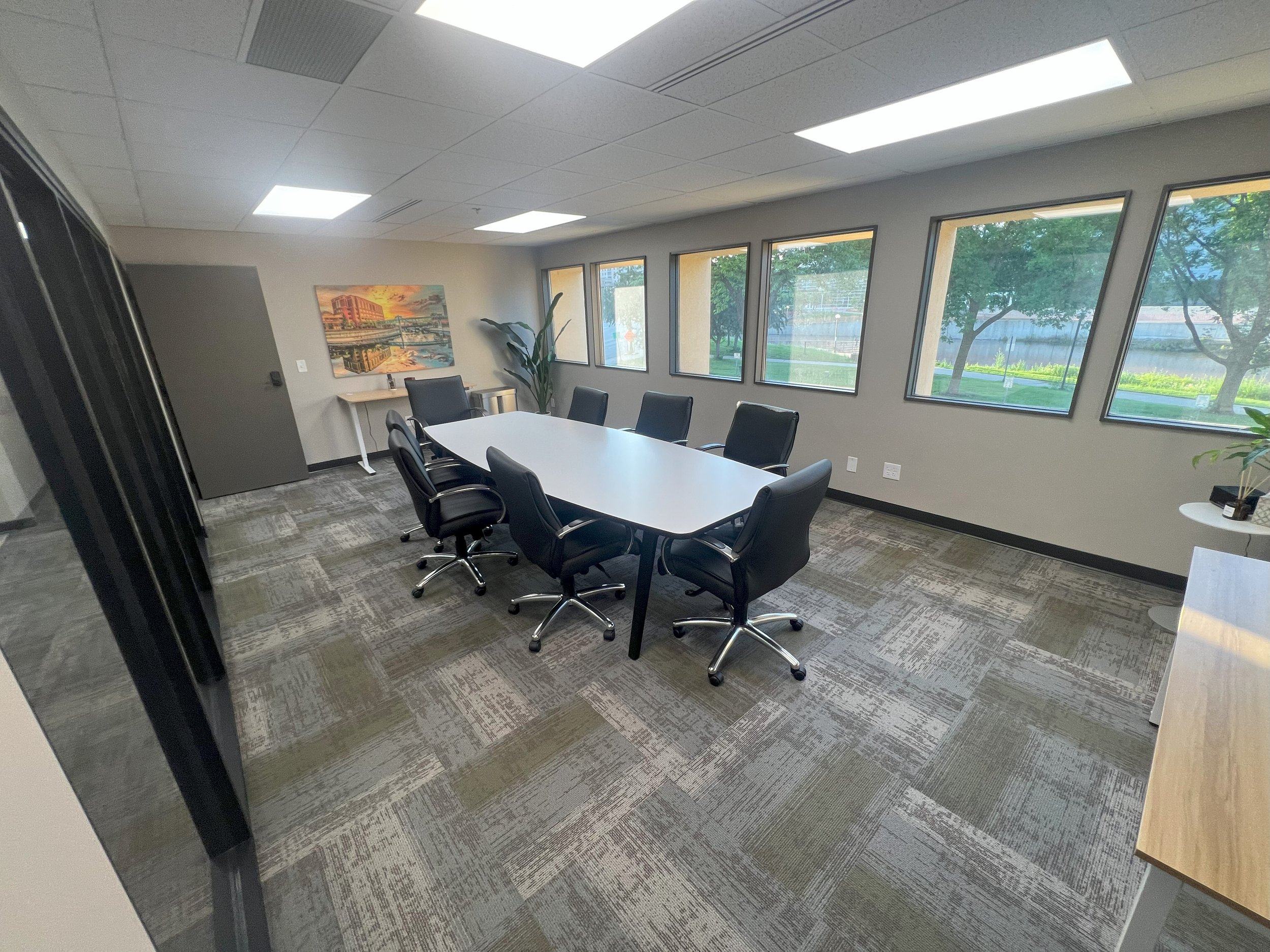 Empty conference room with a long white table surrounded by black chairs, large windows showing trees outside, a colorful painting on the wall, and potted plants.
