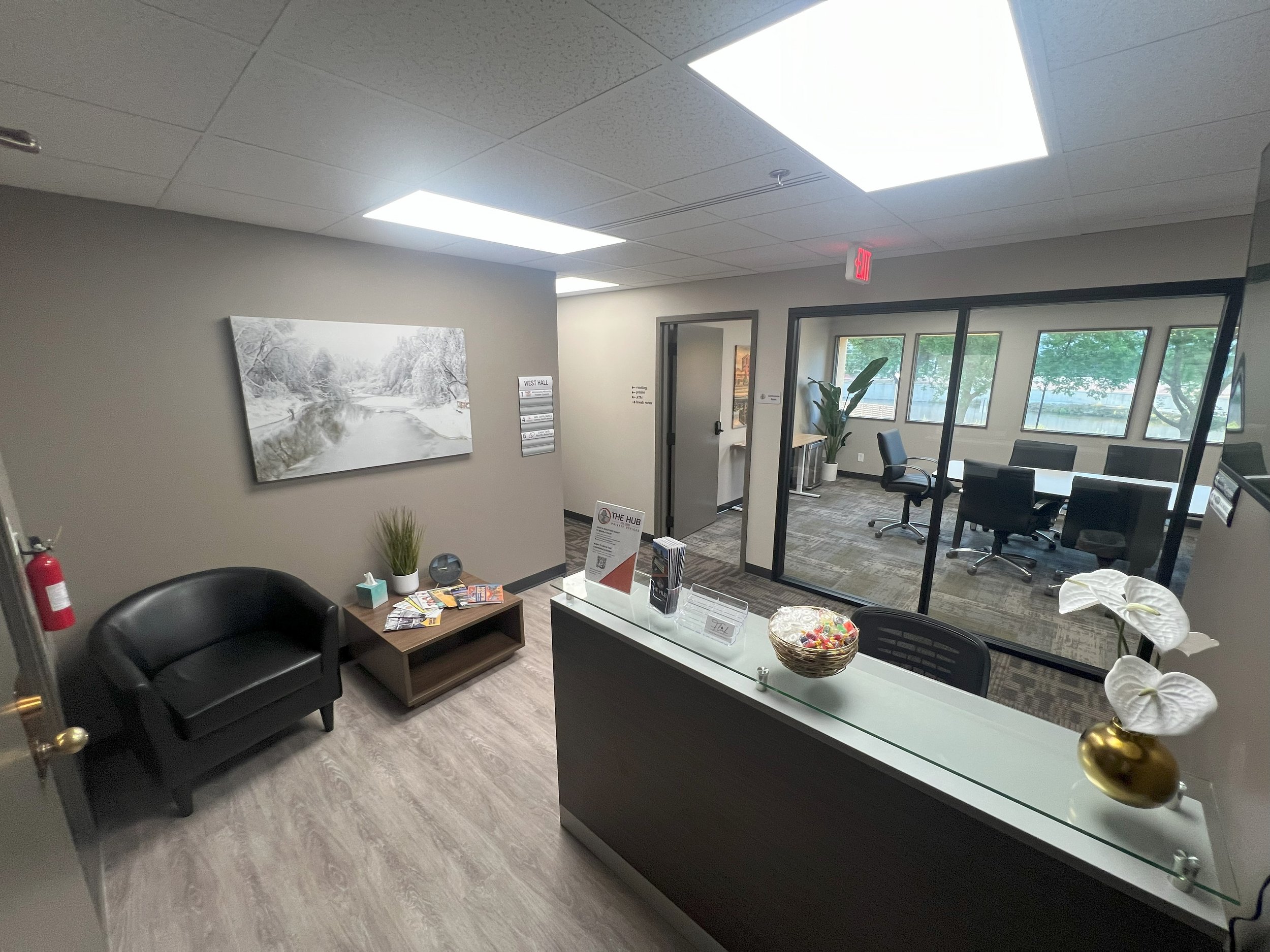 Office reception area with a black sofa, small wooden table, and a reception desk with a bowl of candies. There is a large painting of a snowy landscape on the wall, potted plants, chairs inside a conference room with glass walls, and windows overloo
