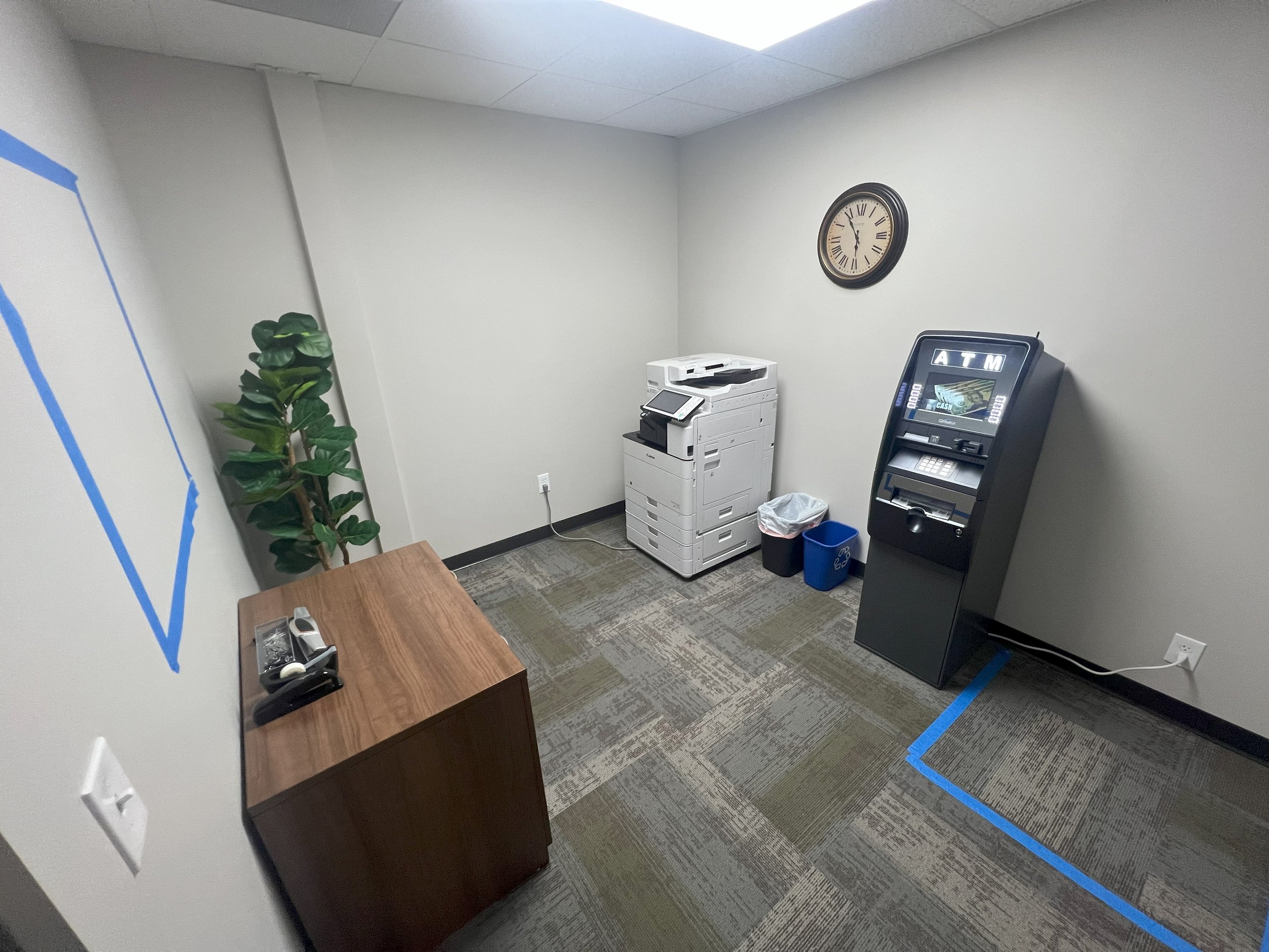Small office room with a wooden desk, potted plant, wall clock, and an ATM machine, with a copier on the corner and two waste bins, one black and one blue. The floor is carpeted with a multicolored pattern, and blue painter's tape marks a section on