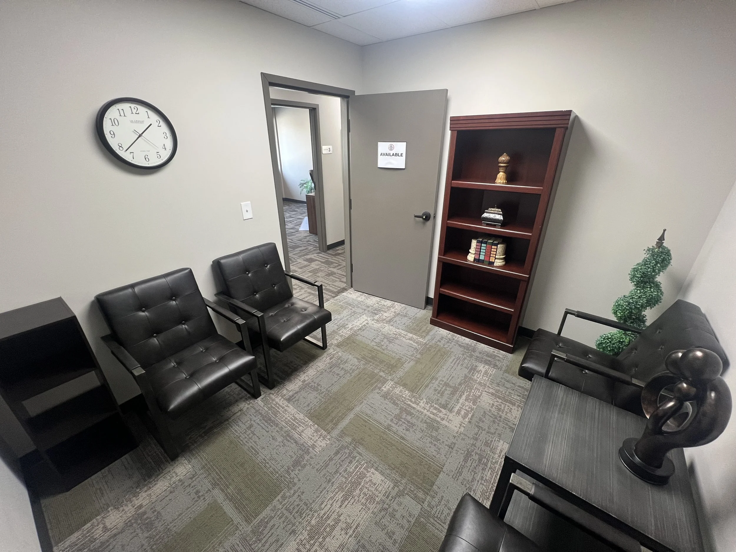 Empty waiting room with four black chairs, a wooden bookshelf, a wall clock, and a door with an 'Available' sign.