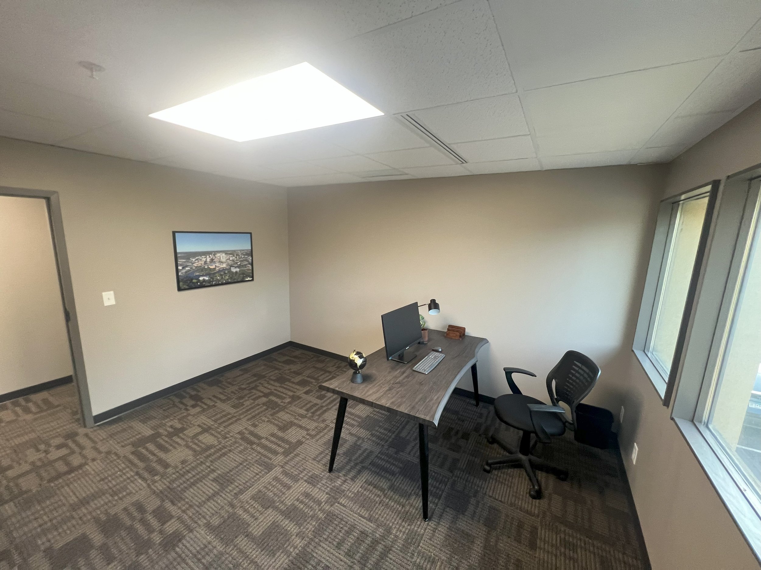 Empty office space with a desk, computer, office chair, and a window, with beige walls and patterned carpet.