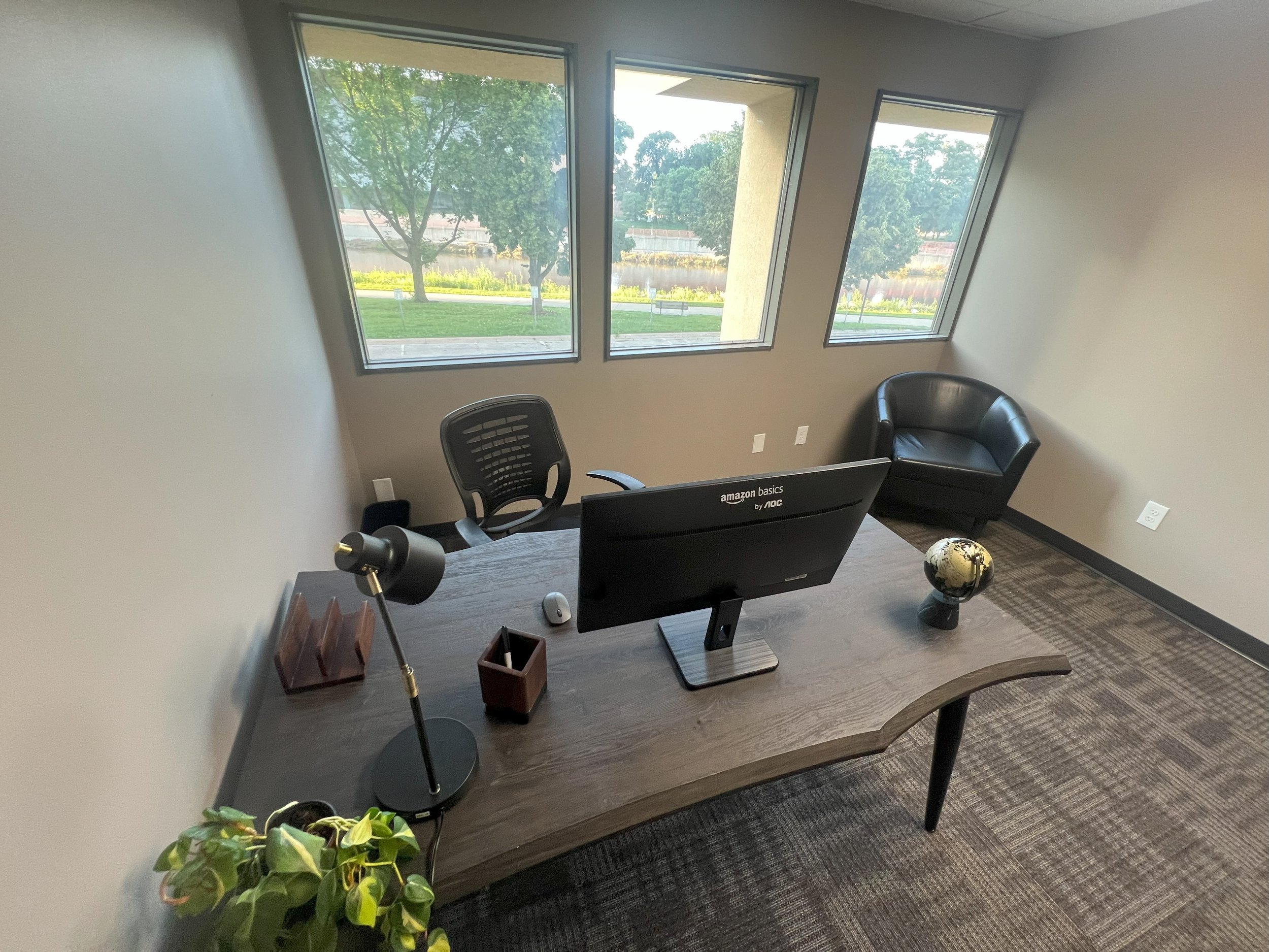Office with a wooden desk, computer monitor, black leather chair, black armchair, globe, desk lamp, and window view of trees and grass.