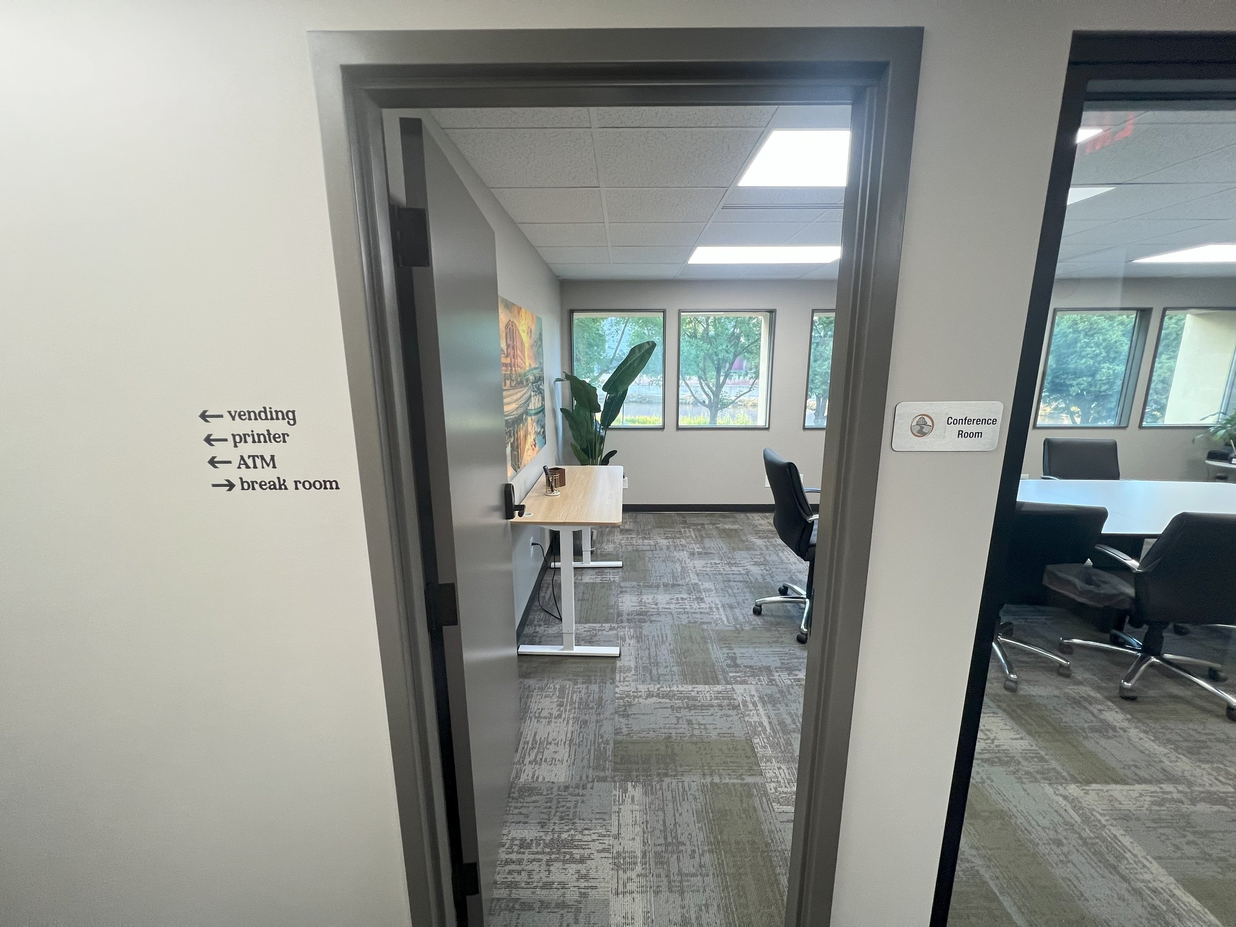 Office conference room with large windows, a white table, black chairs, a potted plant, wall decorations, and signs indicating directions to vending, printer, ATM, and break room.