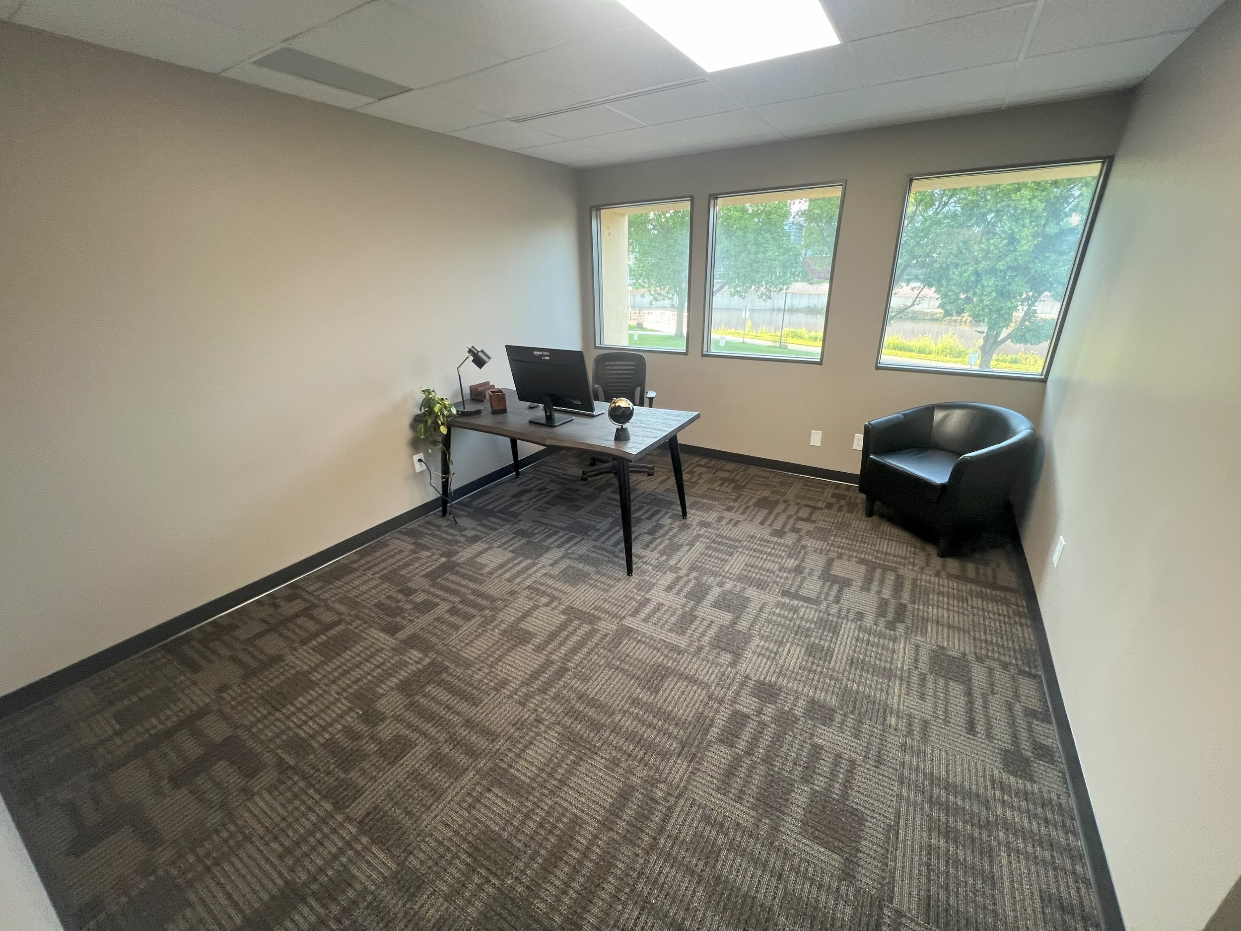 Empty office with a desk, a computer, a black leather chair, and three windows showing greenery outside.