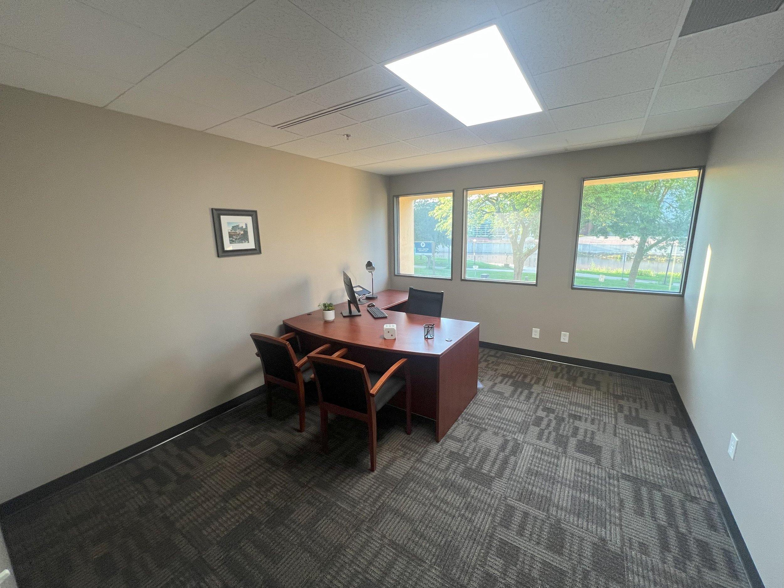 Empty office with a desk, two chairs, a computer, and large windows showing trees and a park outside.