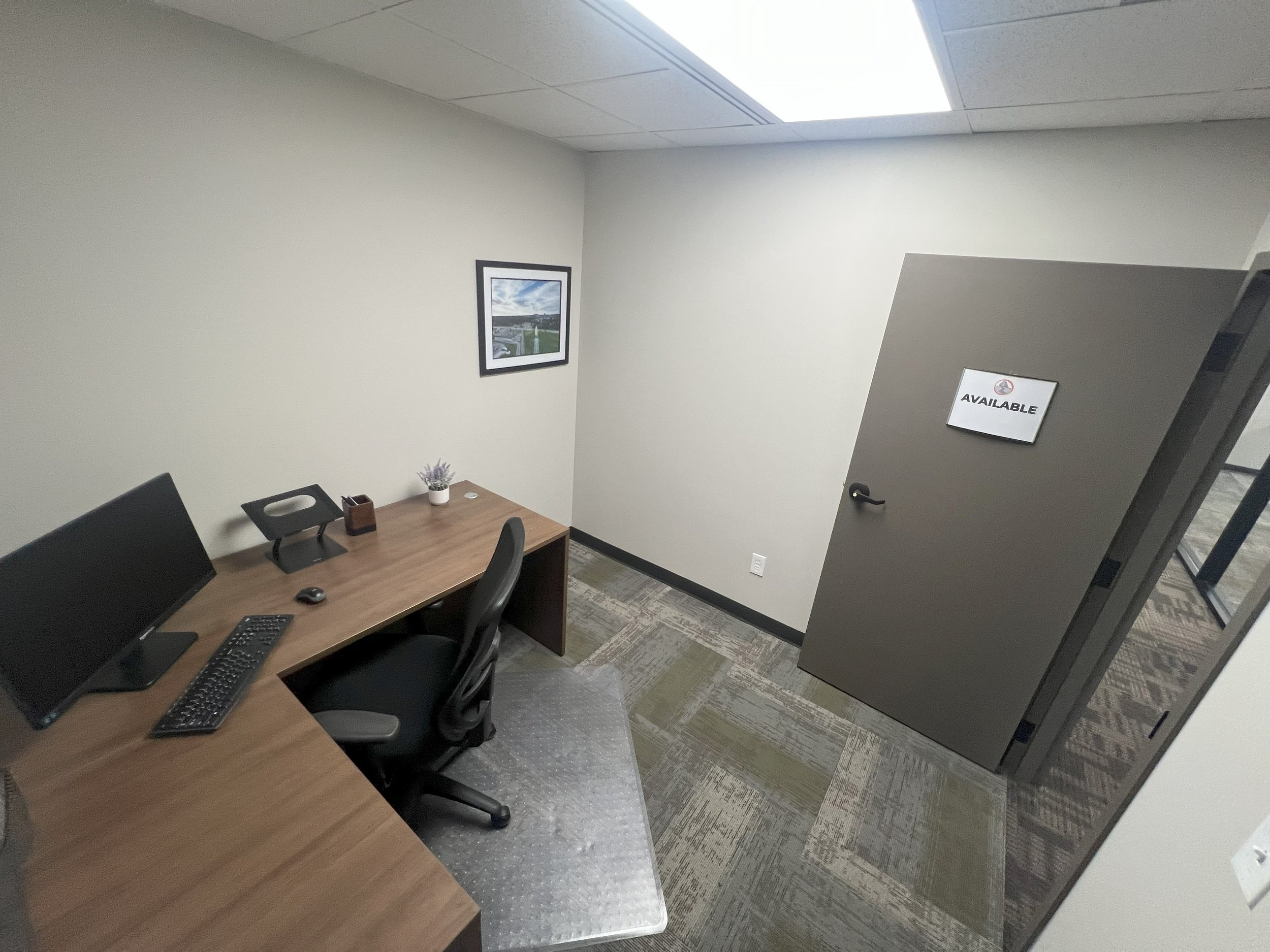 Empty office with desk, computer monitor, chair, picture on wall, and a door with a sign that says 'Available'
