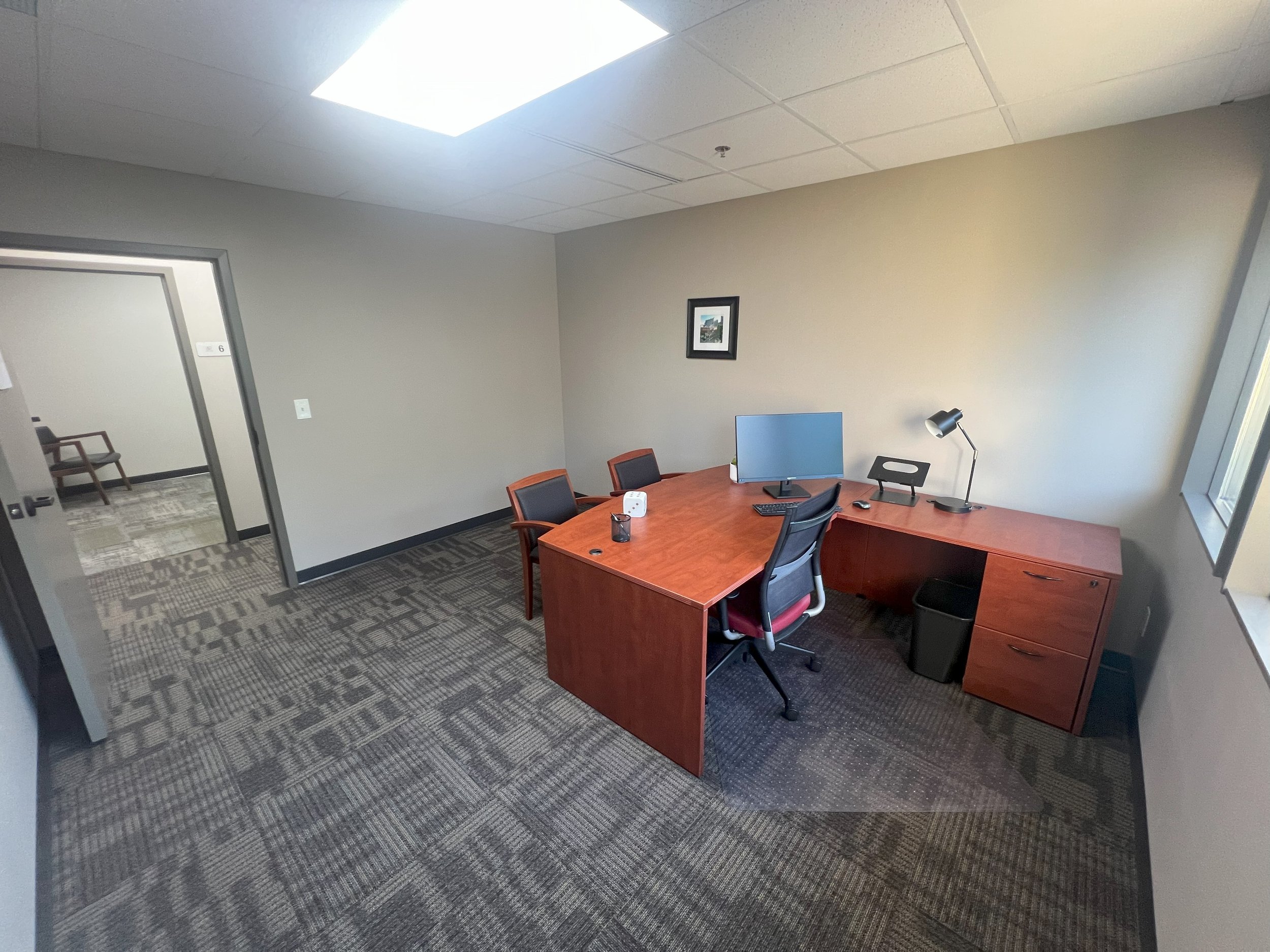 Empty office with a wooden desk, a black office chair, a computer monitor, desk lamp, and two chairs against a beige wall with framed pictures. Office has a patterned carpet and a window on the right side.