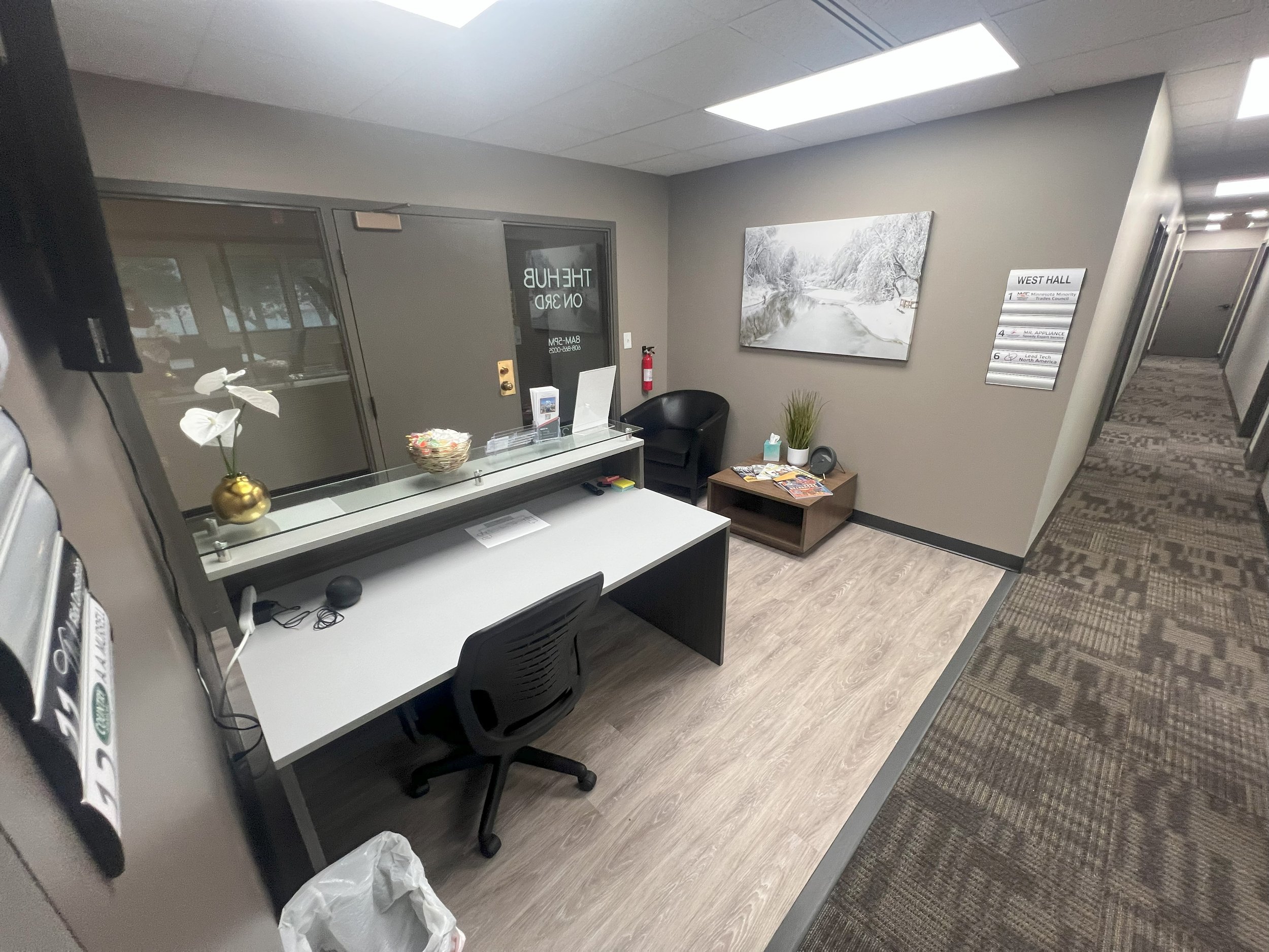 Reception area with a desk, a black chair, a small table with magazines, a potted plant, and a large landscape photograph on the wall, inside a building with gray walls and patterned carpet.
