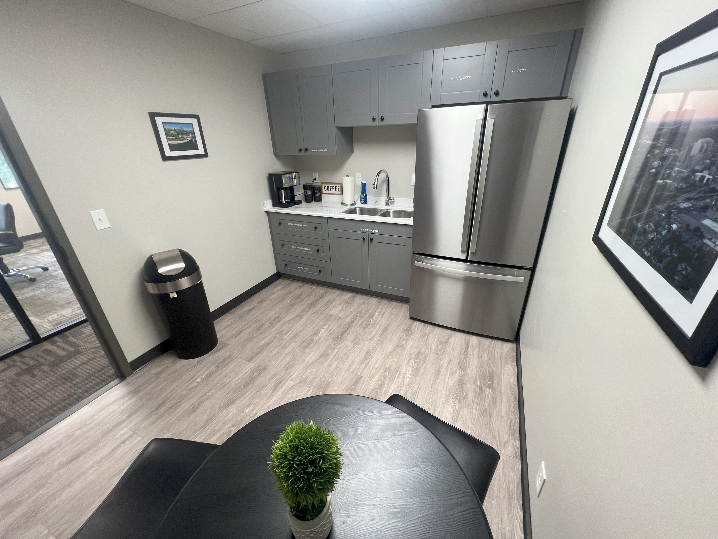 Small kitchen area with gray cabinets, stainless steel refrigerator, coffee maker, and sink. There is a black trash can and a round black table with a small green plant. Two framed pictures hang on the walls, and a sliding glass door leads to an outd