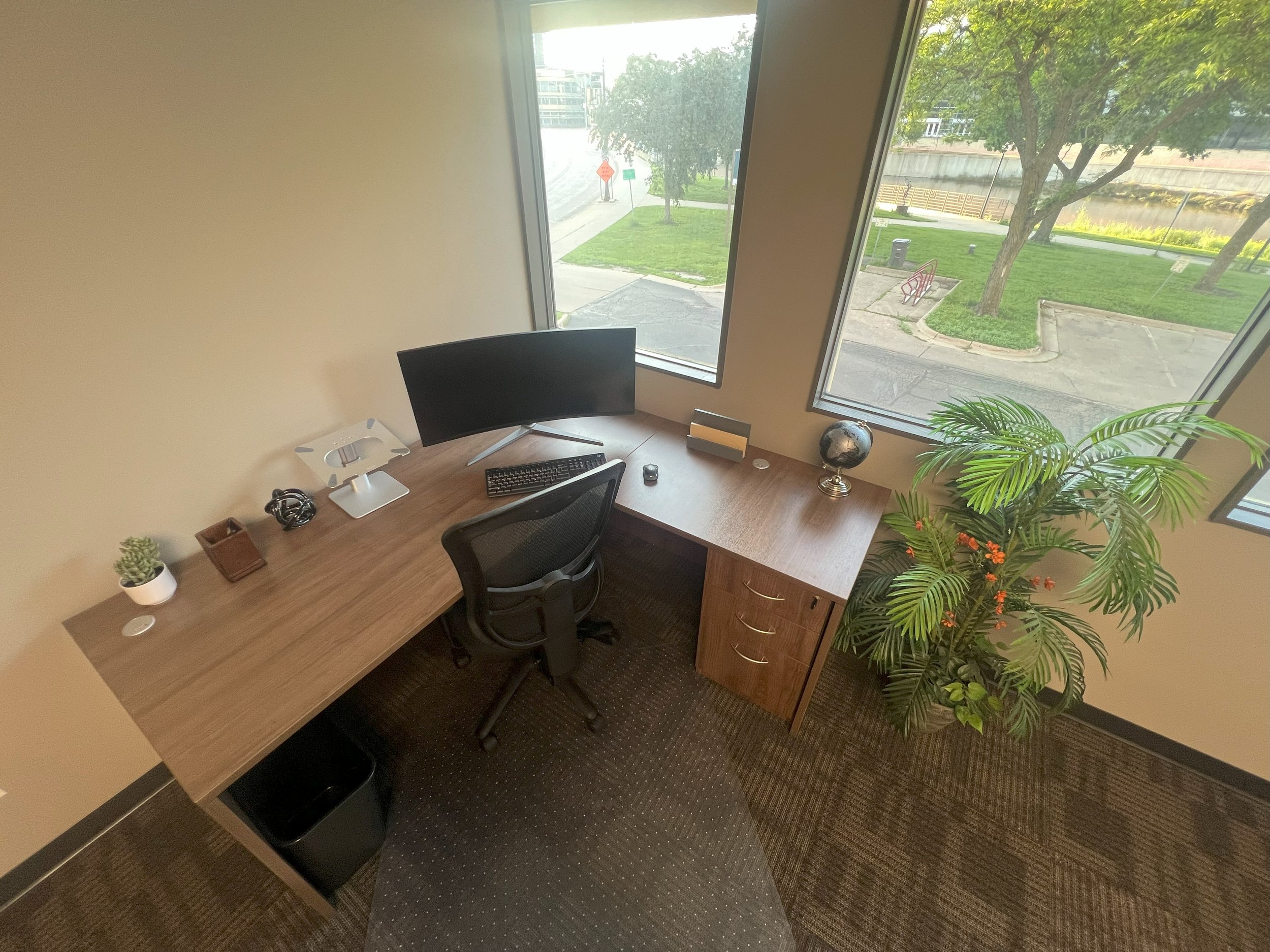 A small office space with a wooden desk, black office chair, and a computer monitor near large corner windows. The desk holds a small white plant, a small brown pot, headphones, a globe, a speaker, and a small object. A large potted plant is next to