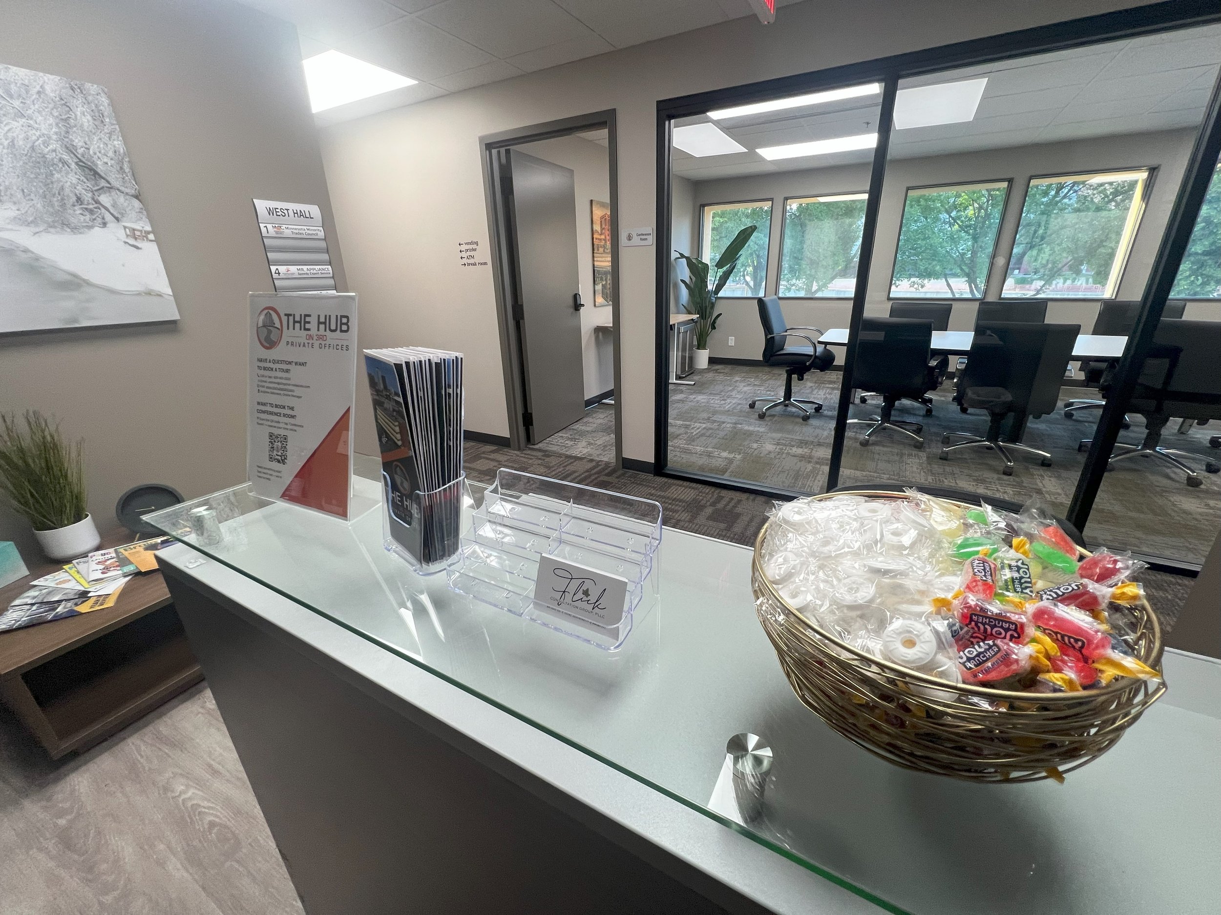 Reception desk with a basket of candy, informational brochures, and a sign for private offices, in an office lobby with conference room visible through glass walls.