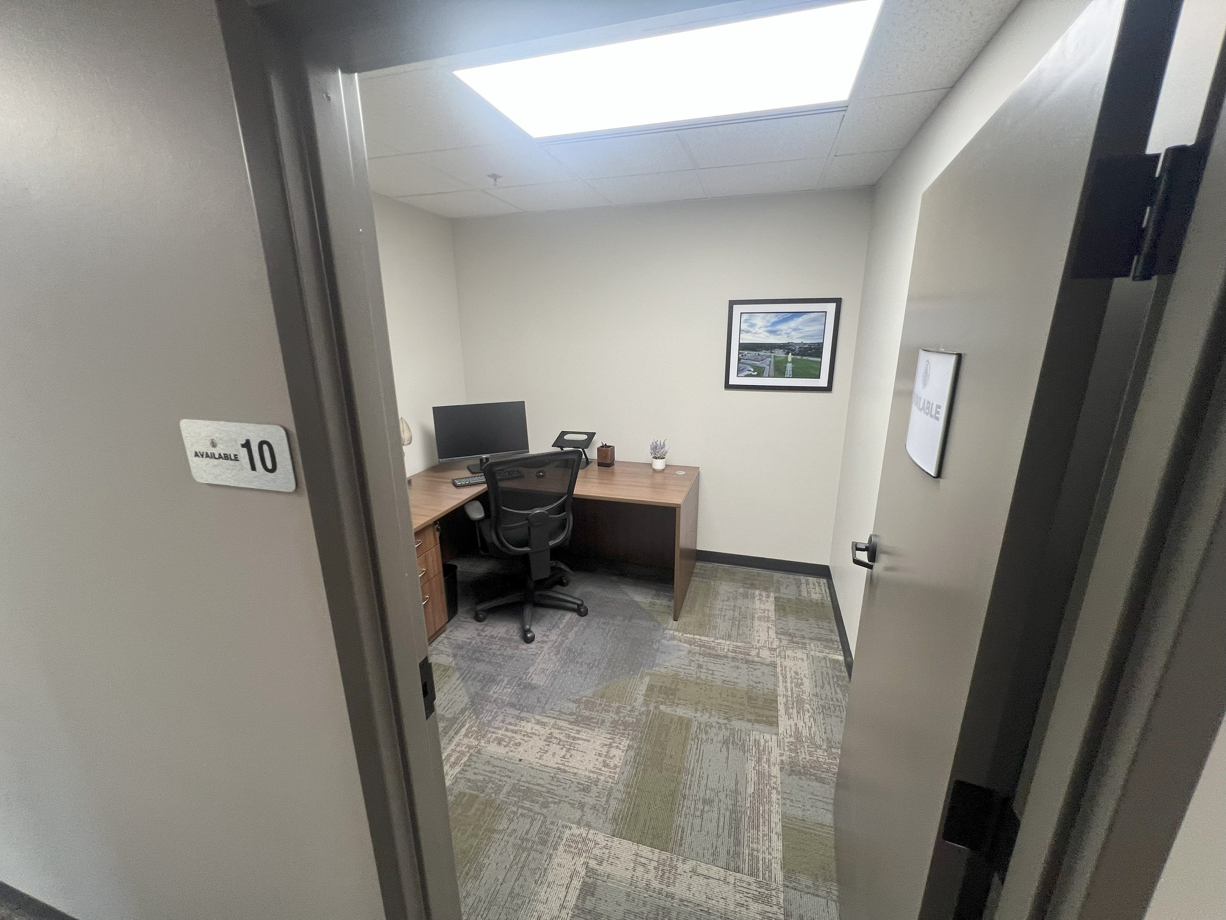 Empty office cubicle with a desk, a computer monitor, a chair, a small plant, a framed picture on the wall, and office supplies, viewed through an open door with a sign indicating room 10 is available.