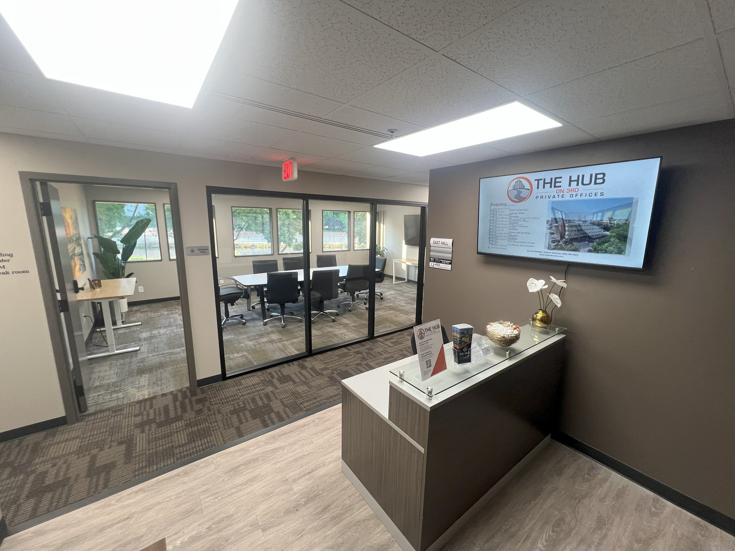 Office reception area with a glass-walled conference room, a large wall-mounted screen, and a small desk with decorative items.