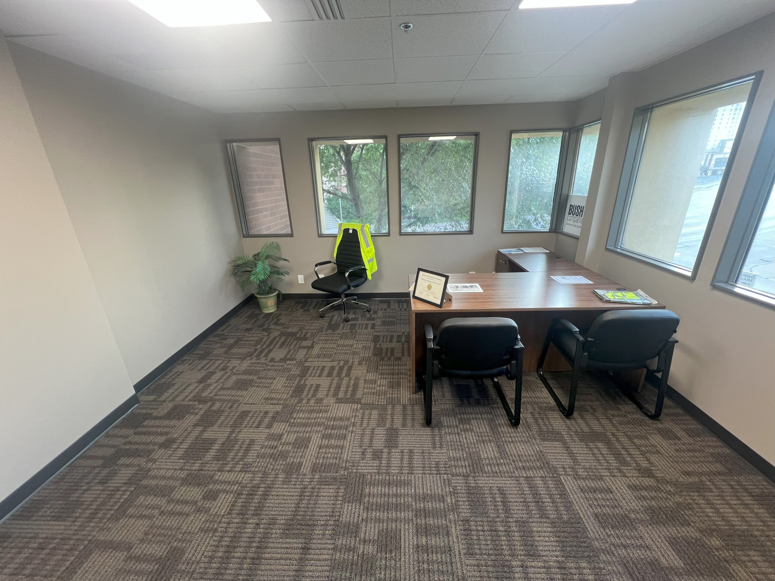 An empty office with a wooden desk, two black chairs, a black office chair with a neon yellow safety vest hanging on the back, a small table with paperwork, a framed certificate, a potted plant, and a window view of trees and buildings.