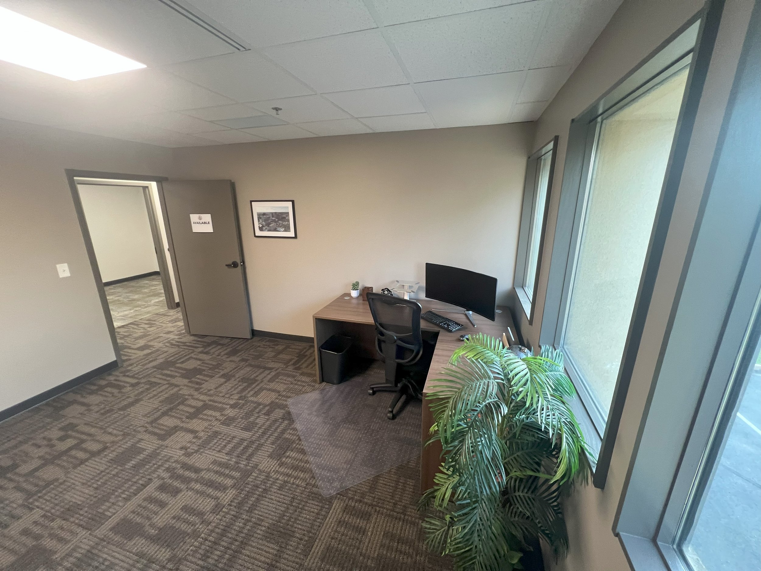 Office with beige walls, a desk with a computer, black office chair, a potted plant near a window, and an open door leading to another room.