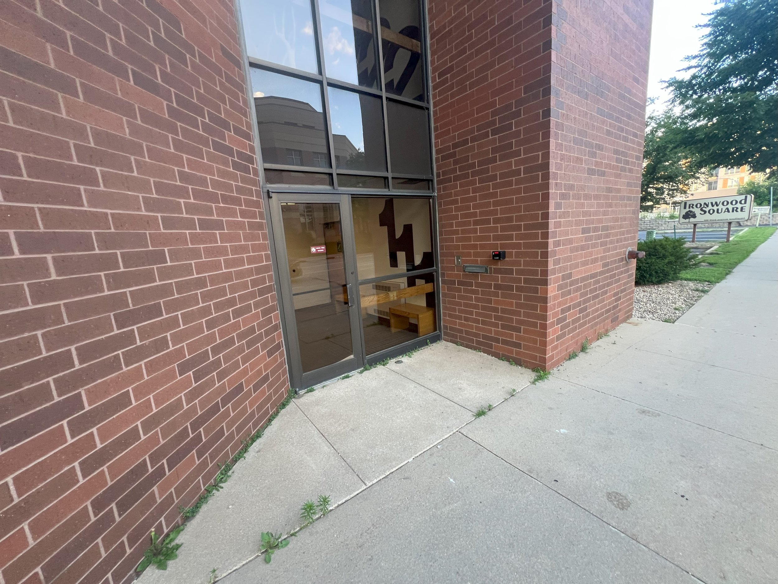 Shiny glass door of a brick building with a bench inside visible through the glass. A sidewalk and a sign that reads 'Ironwood Square' along with some greenery are outside.
