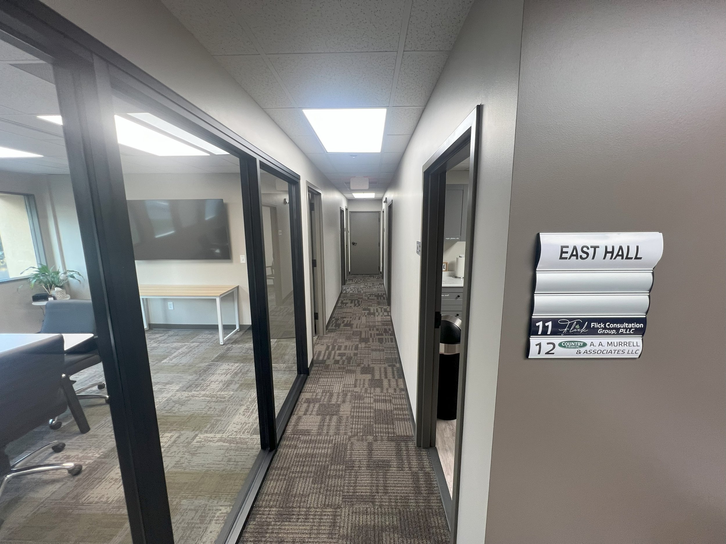 An office corridor with glass-walled conference rooms and doors, gray walls, patterned carpet, and a sign indicating rooms 11 and 12 for East Hall, with workplace furniture visible through the glass.