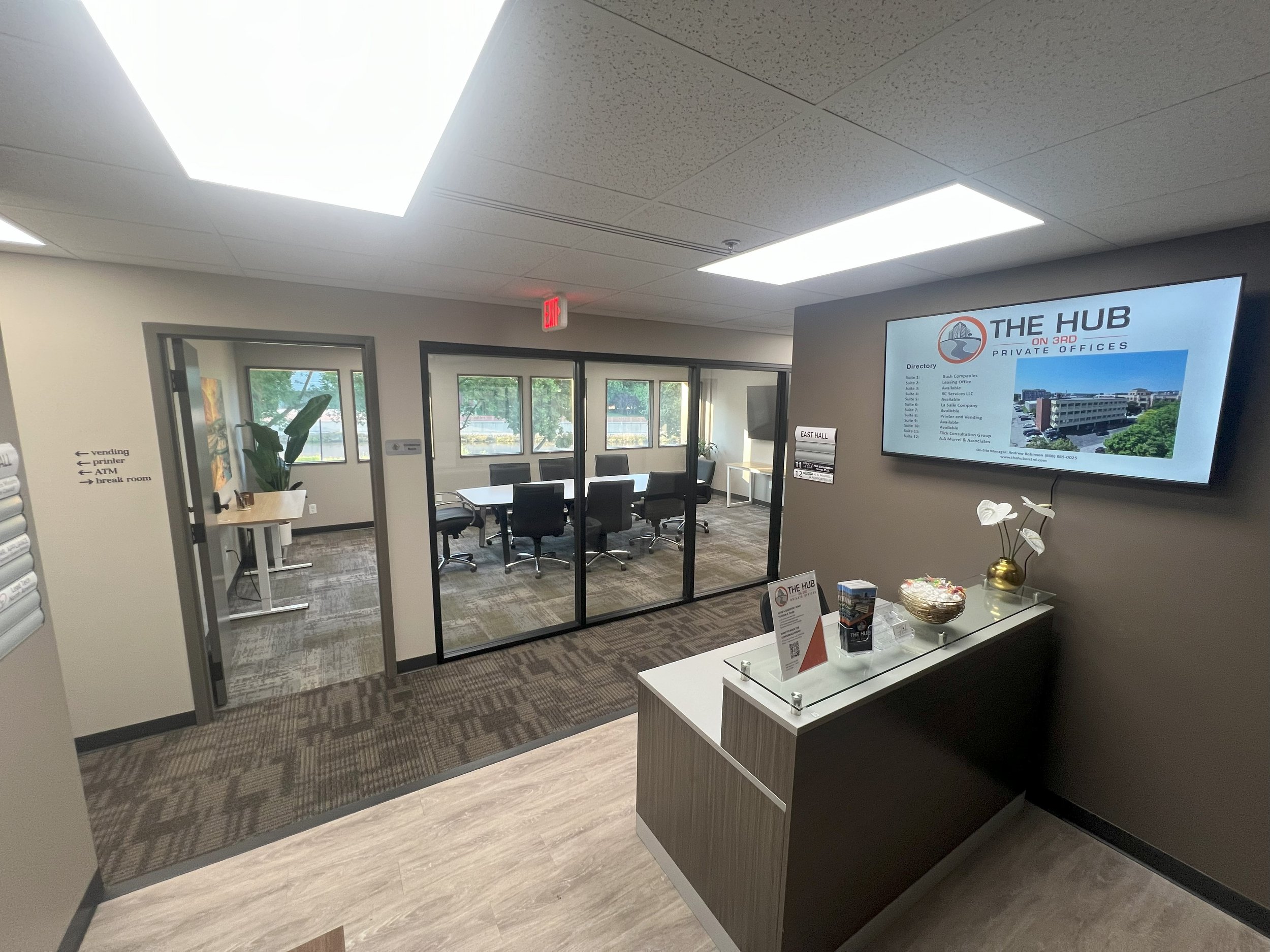 Reception area with a wall-mounted display screen showing 'The Hub on 3rd Private Offices' and a directory list, a table with informational brochures, behind glass conference rooms with chairs, a large window, and signs indicating vending, printer, A