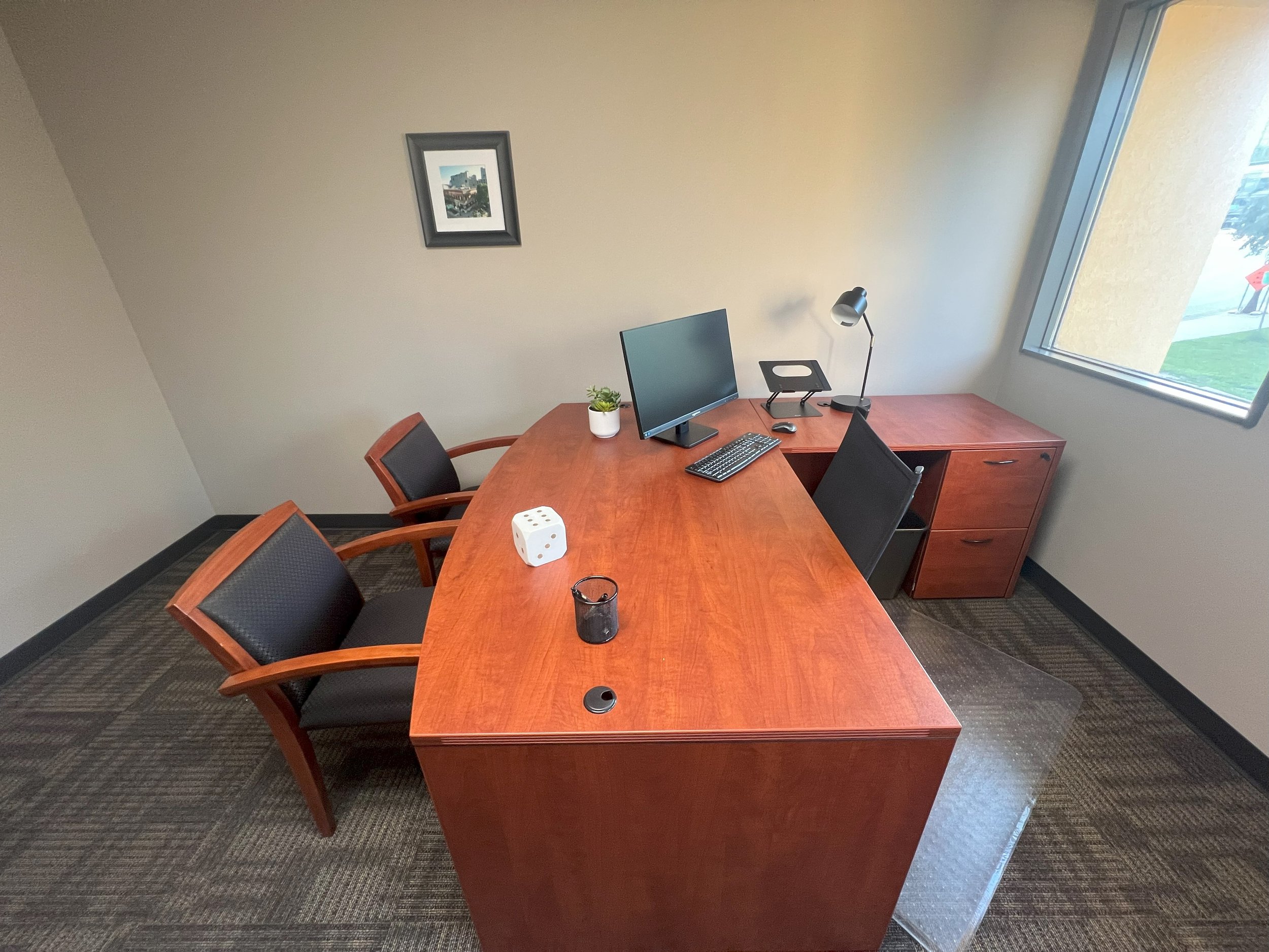 Empty office with a wooden desk, three chairs, a computer monitor, a keyboard, a mouse, a desk lamp, a small plant, a die, a black pen holder, and a desk organizer. There is a framed picture on the wall and a large window to the right.