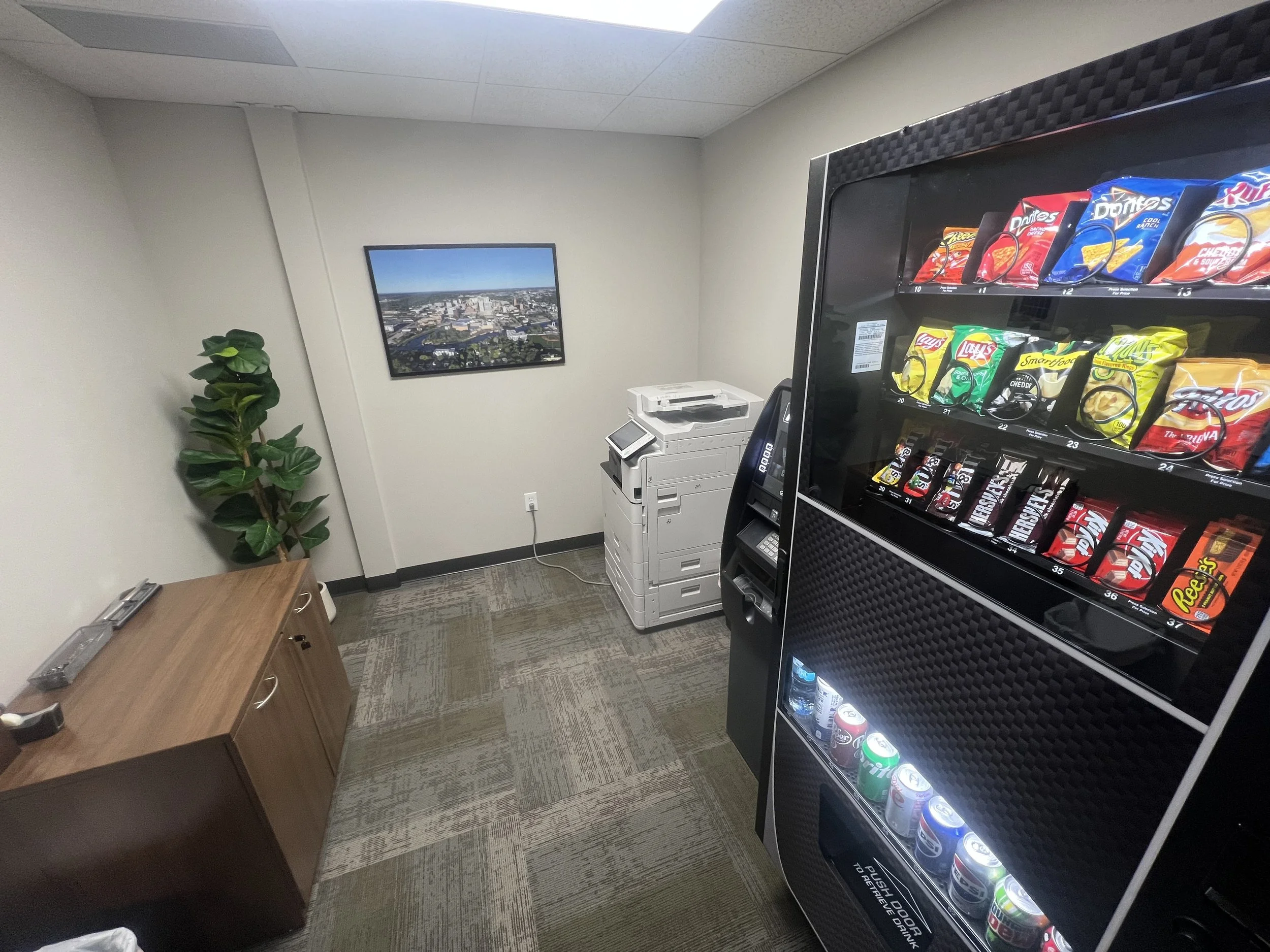 Office break room with vending machine, printer, wooden cabinet, potted plant, and a framed picture of a cityscape on the wall.