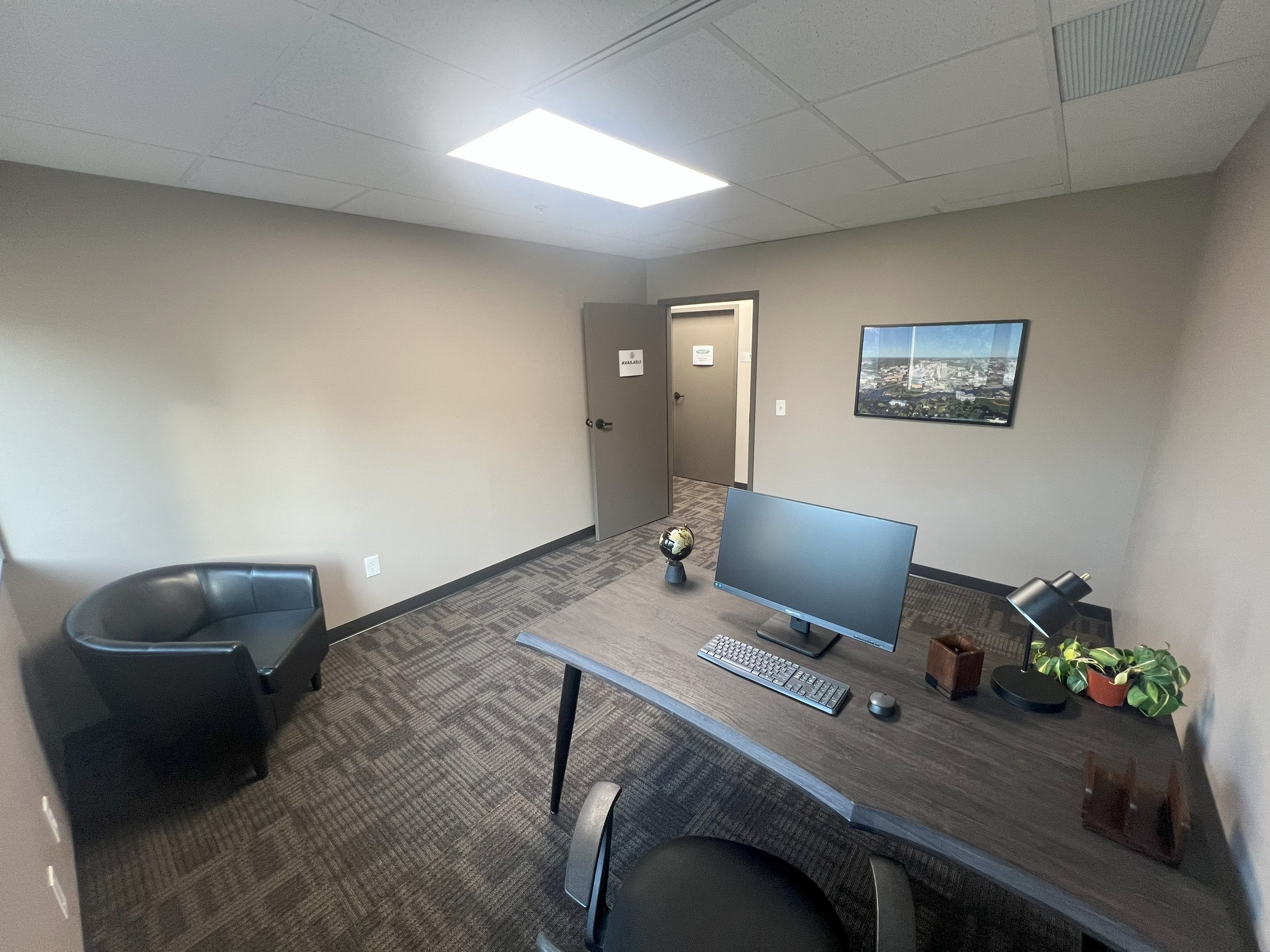An empty office with a desk, computer monitor, keyboard, mouse, a globe, a desk lamp, a potted plant, and two chairs. There is a framed picture on the wall and a door in the background.