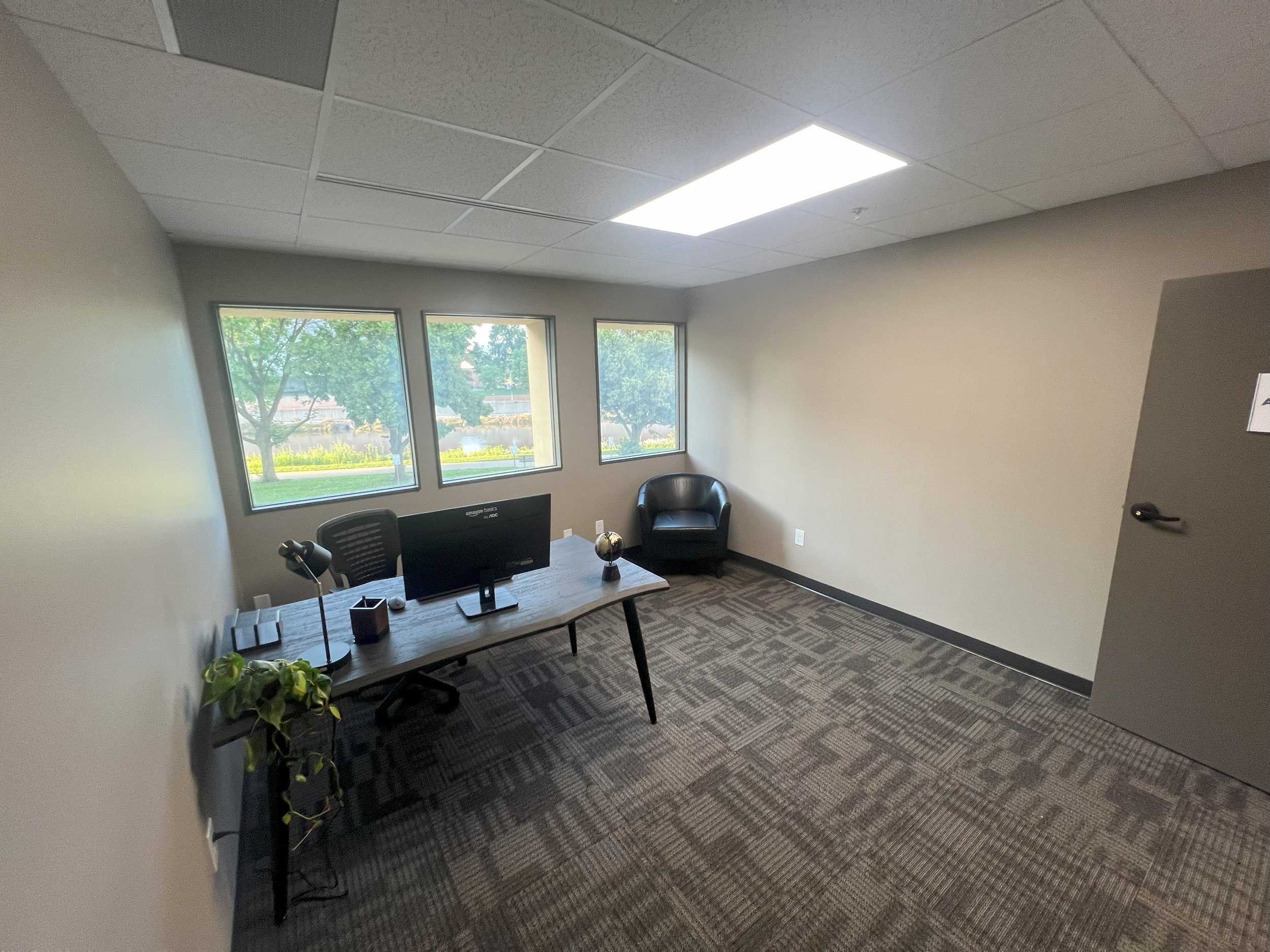 Empty office room with a desk, chair, computer monitor, lamp, small globe, and wall-mounted power strip, with three large windows showing trees outside.