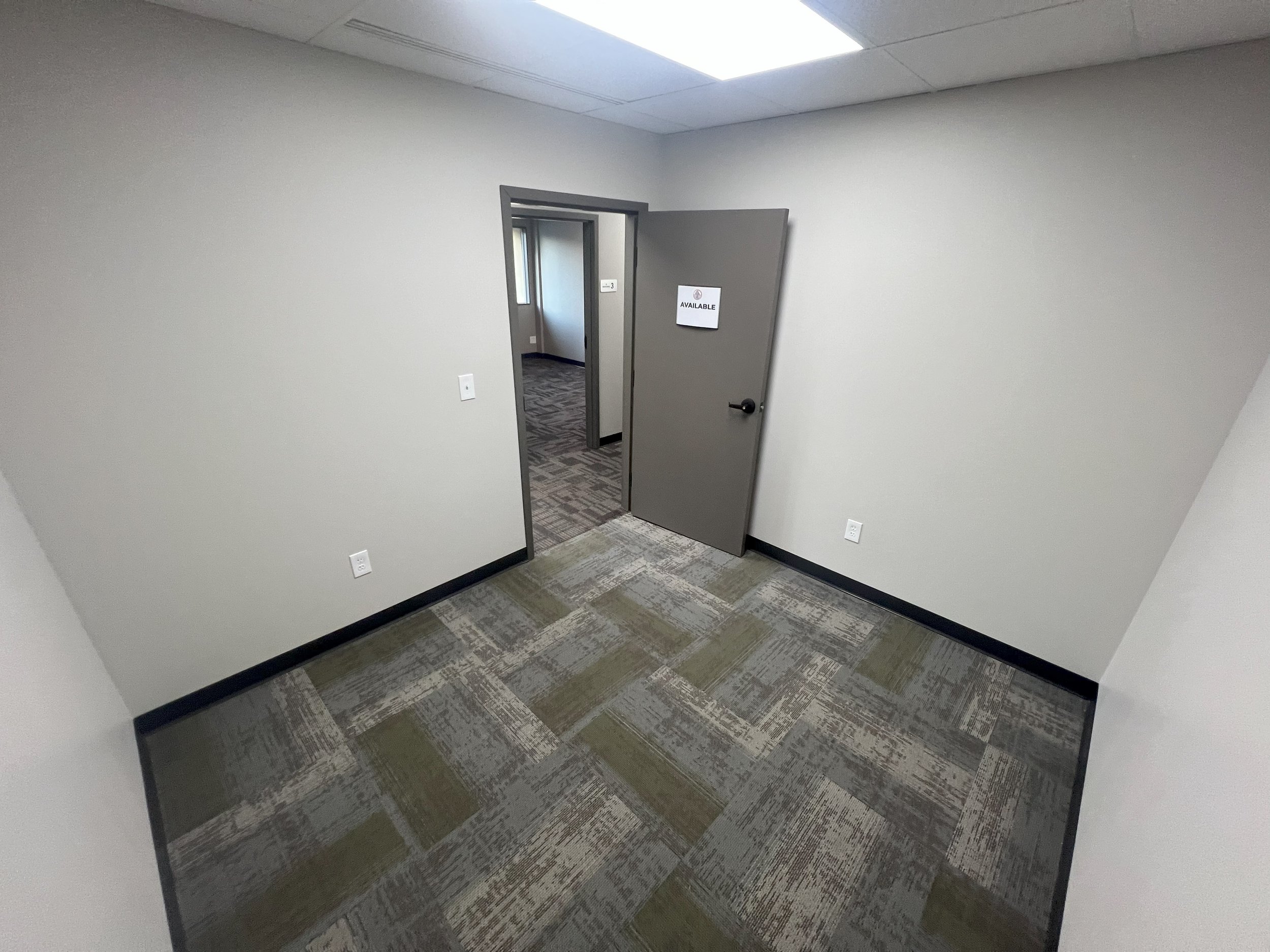 Empty office room with gray walls, a door labeled 'Available,' and carpeted floors.