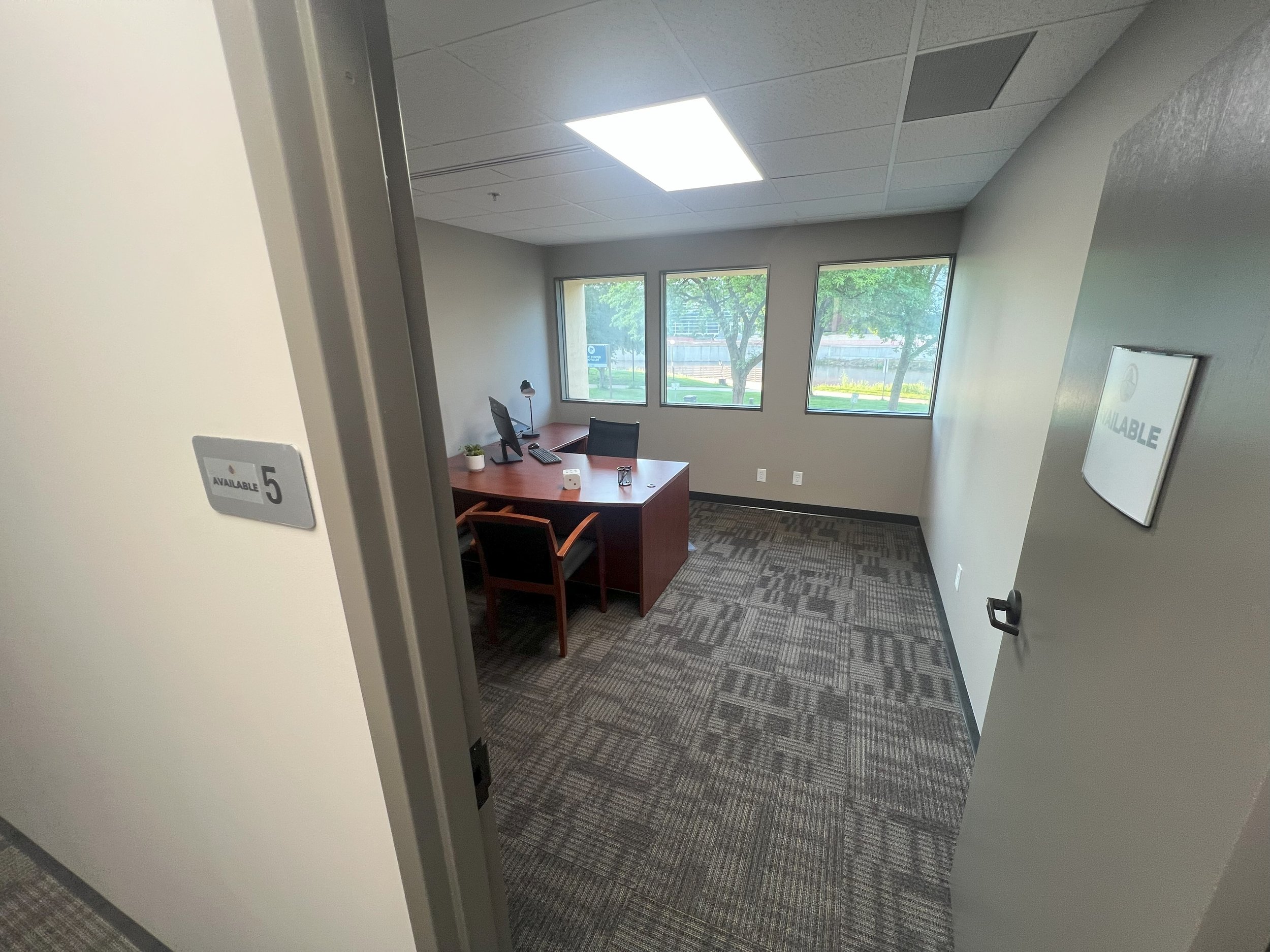 Empty office with a wooden desk, black office chair, large windows, and gray walls.