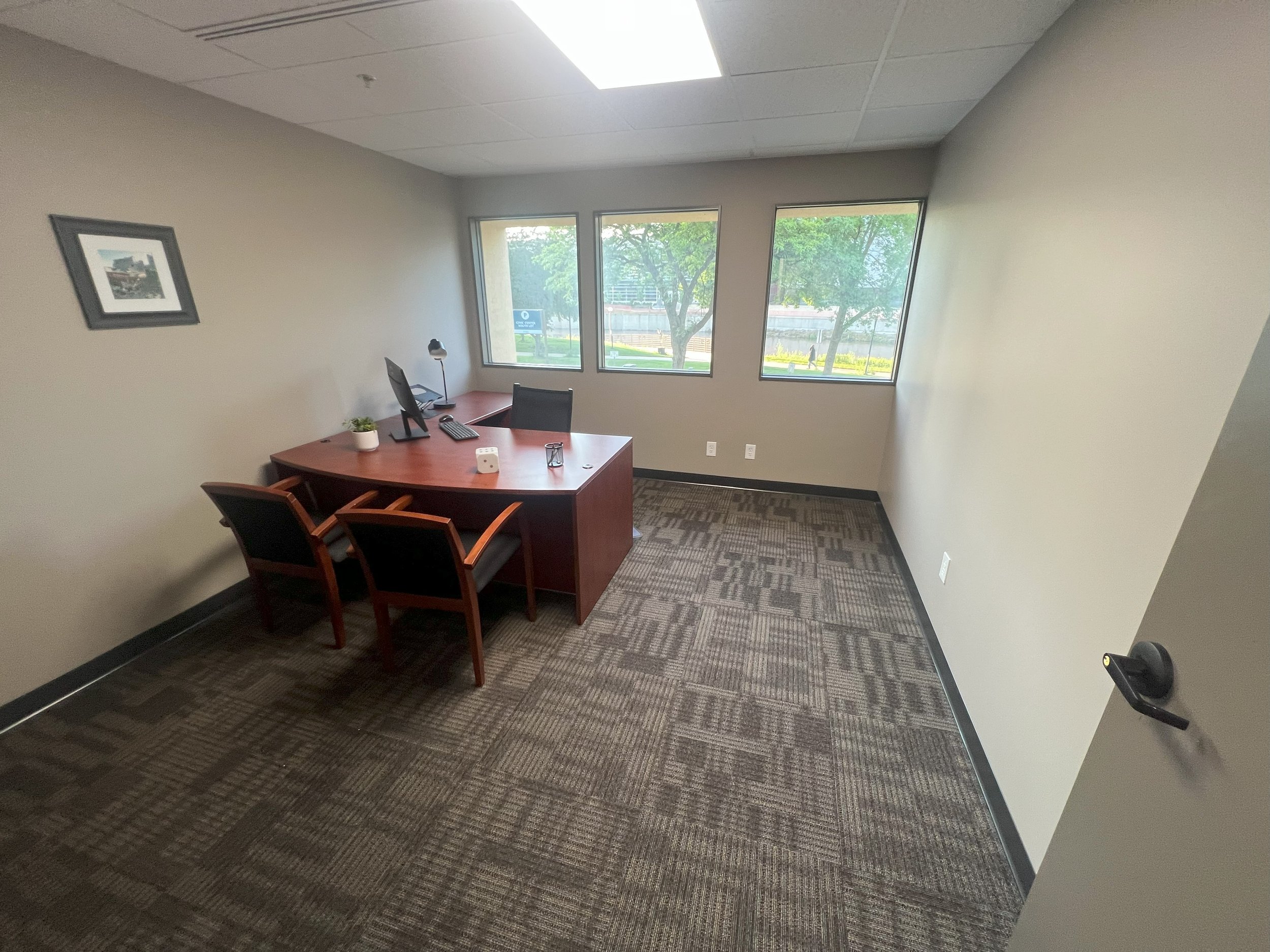 Empty office with a wooden desk, three chairs, a computer, and windows with a view of trees outside.