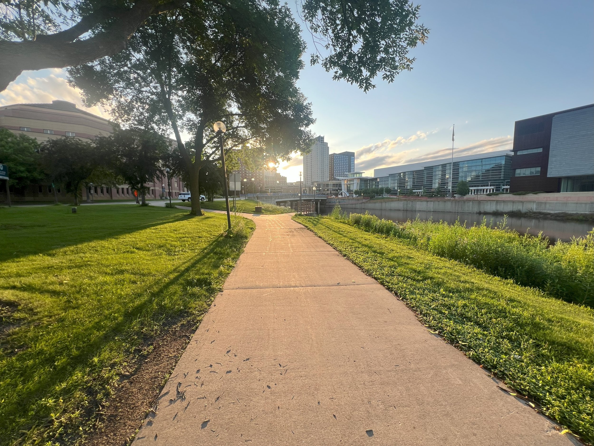 Paved walkway along a grassy park with trees, modern buildings, and water on the right side, under a sunny sky.