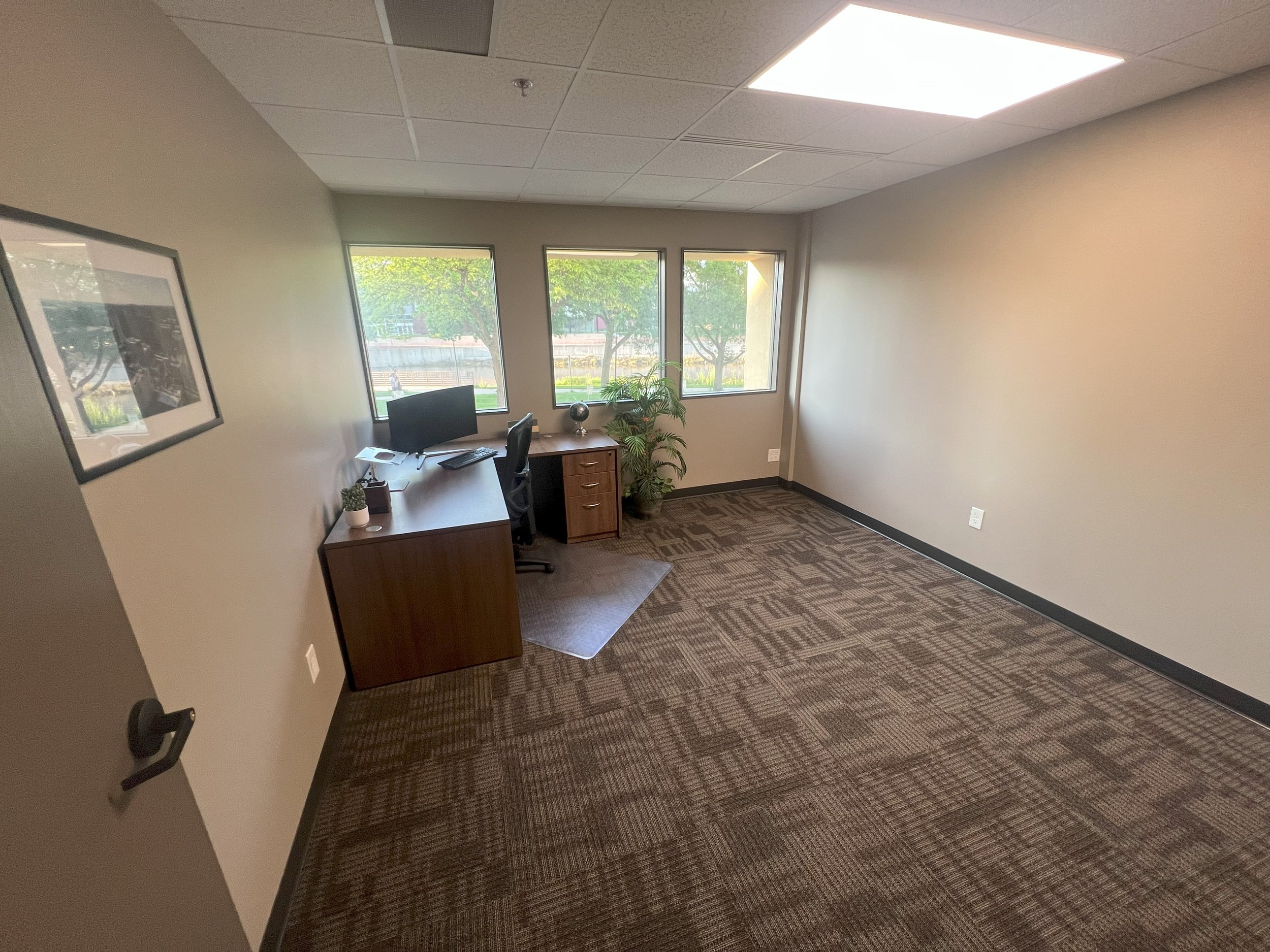 Empty office room with beige walls, large windows, a wooden desk with a computer and chair, a small plant, and a framed picture on the wall.
