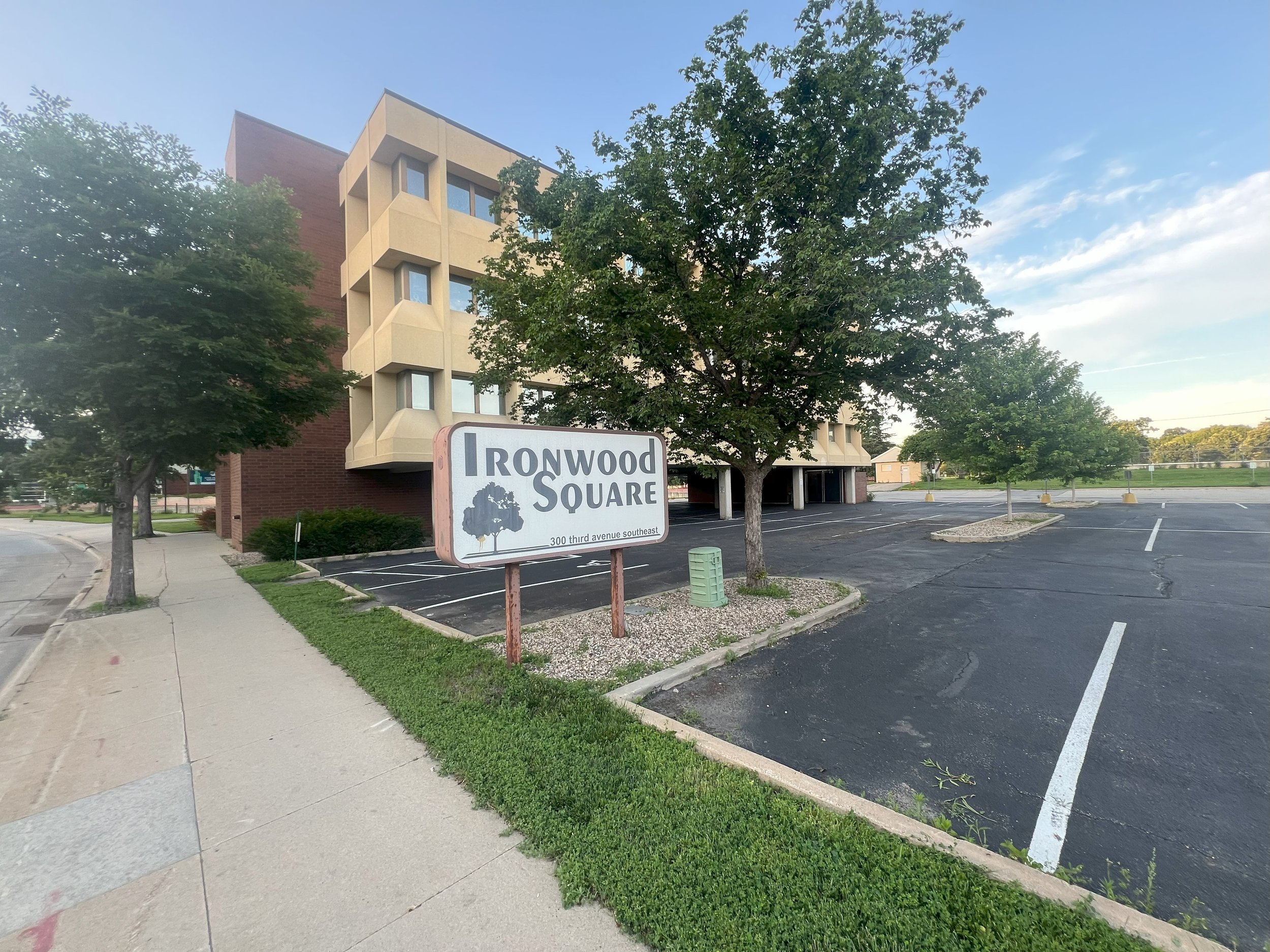 Empty parking lot beside a building with trees, a sign reading 'Ironwood Square' in front, and clear sky.
