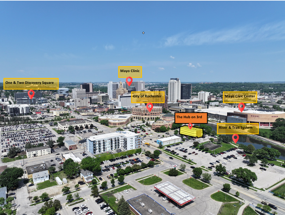 Aerial view of downtown Rochester, New York with labeled landmarks including Mayo Clinic, City of Rochester, Mayo Civic Center, One & Two Discovery Square, The Hub on 3rd, and River & Trail System, under a blue sky with scattered clouds.