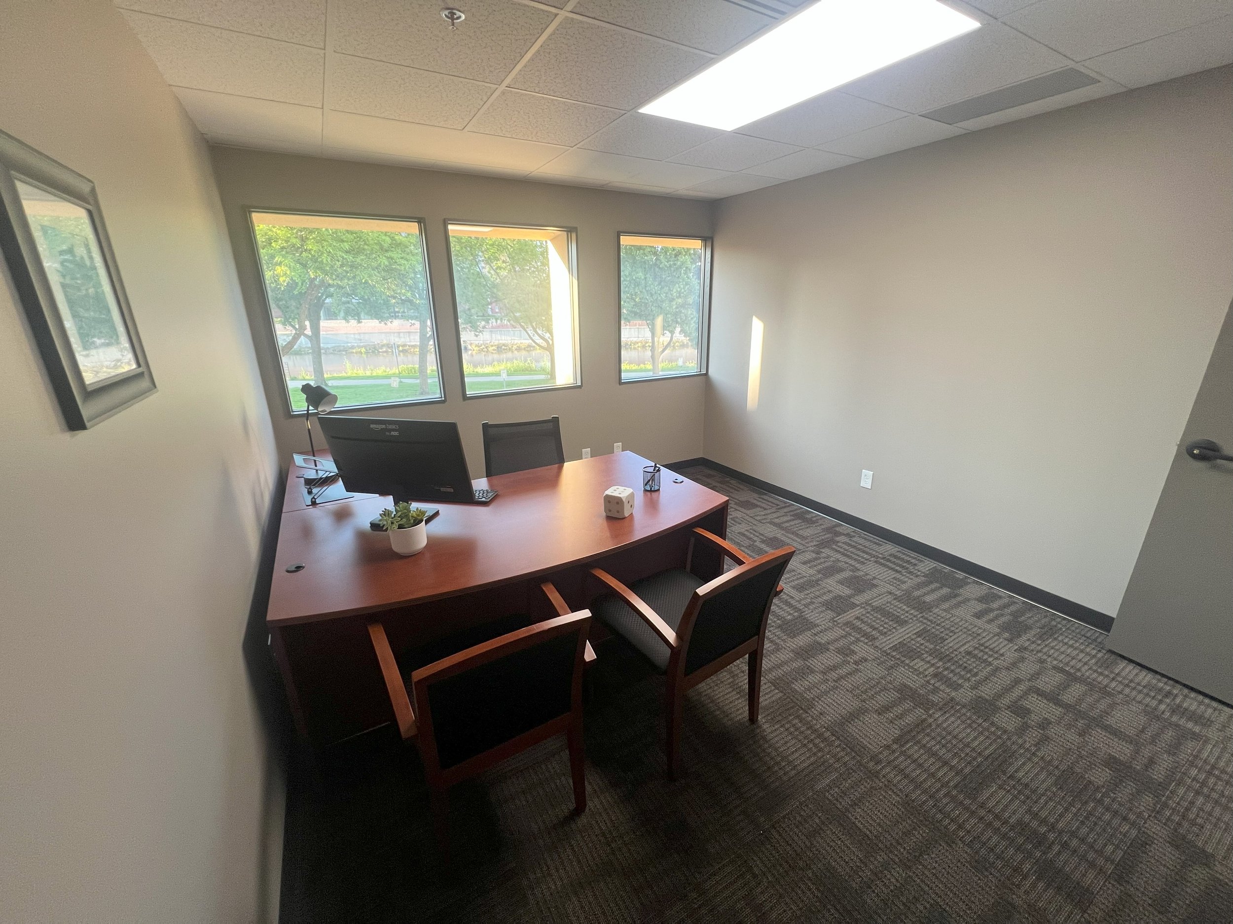 An empty office with a wooden desk, two chairs, a computer monitor, a potted plant, and a window with trees outside.