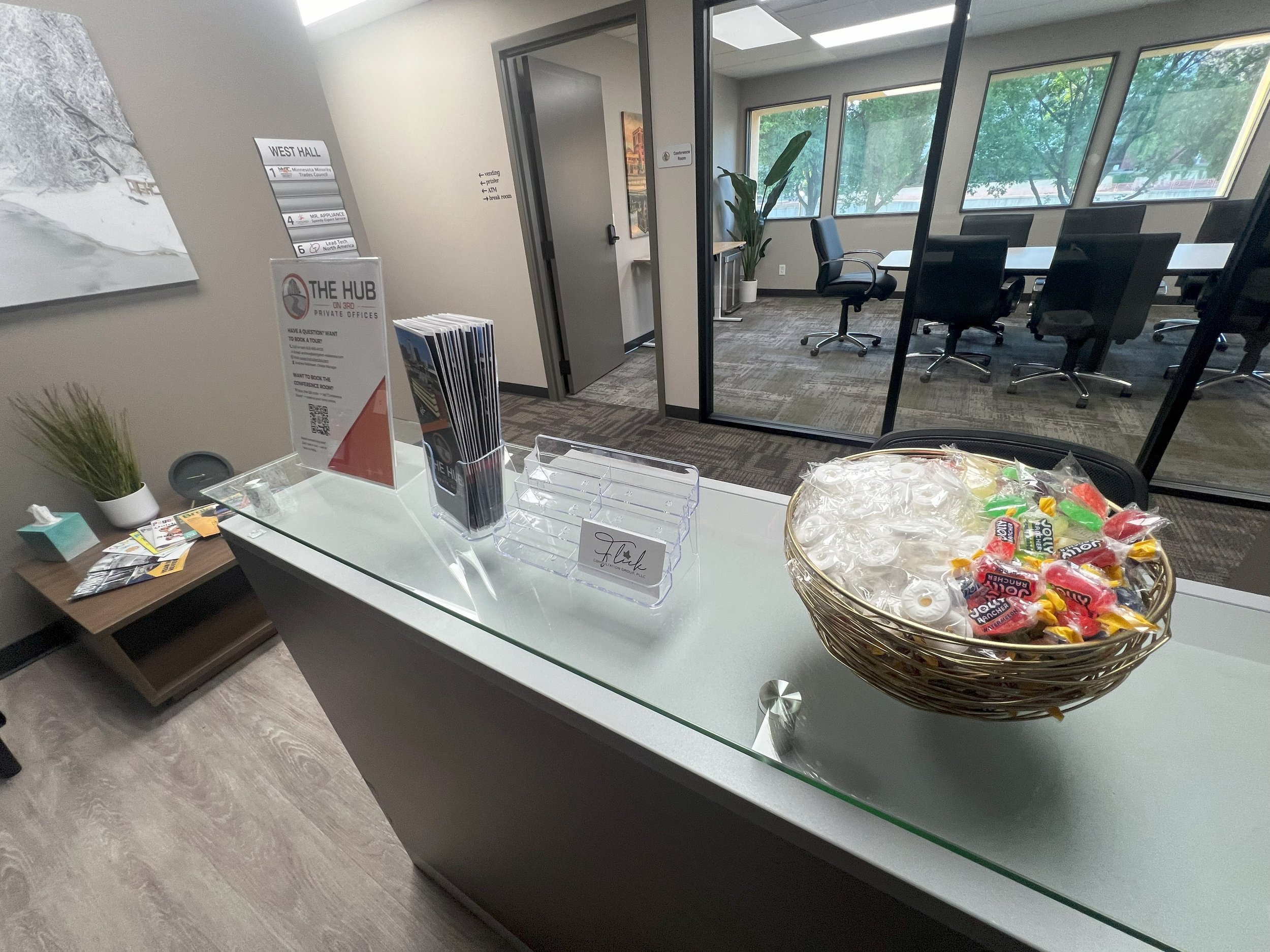 Reception desk with a basket of candies and a clear brochure holder, adjacent to a waiting area and conference room with large windows and office chairs.