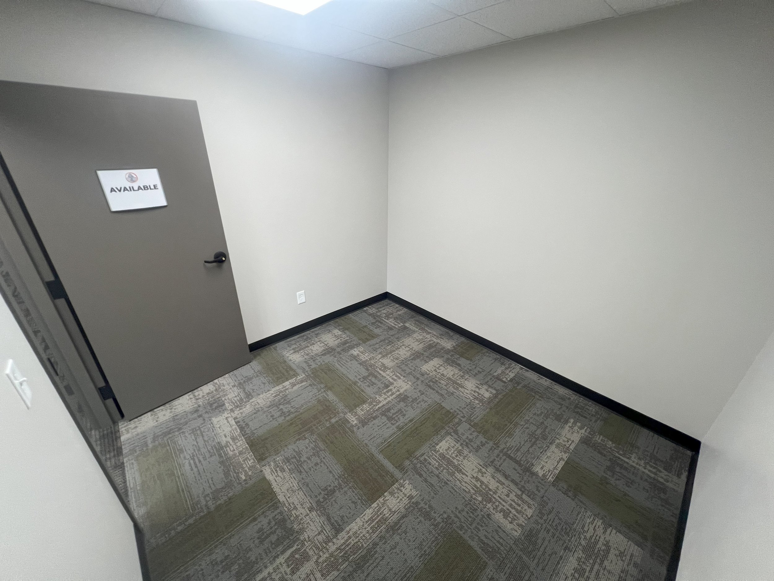 Empty office room with a closed gray door that has a sign reading 'Available' and a black handle, beige walls, and patterned carpet flooring.